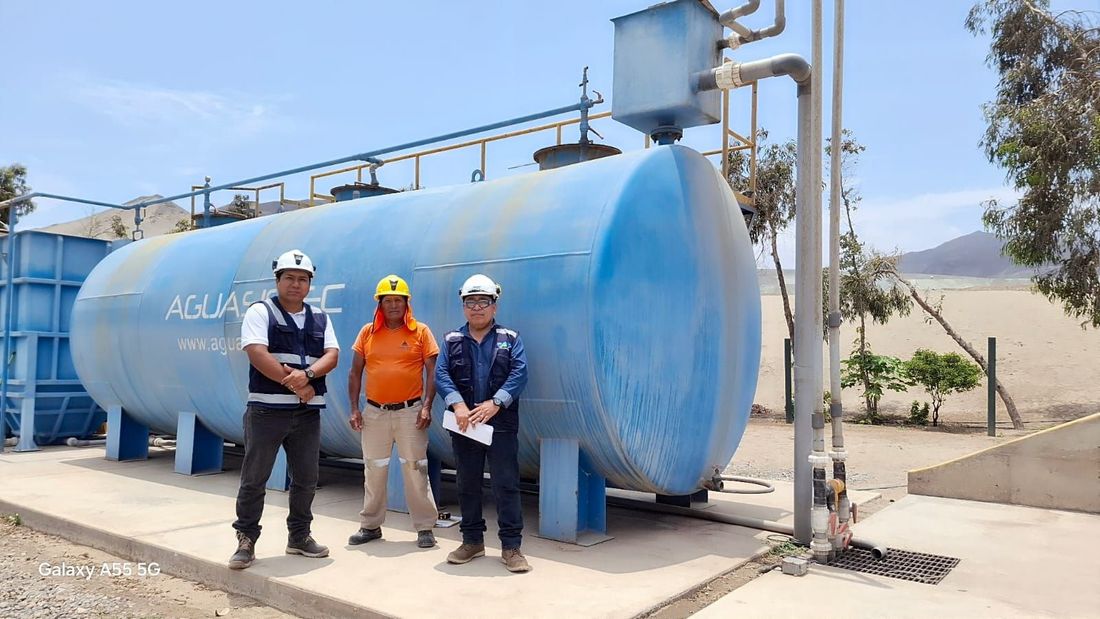 Tres trabajadores con cascos de seguridad de pie frente a un gran tanque industrial azul al aire libre.