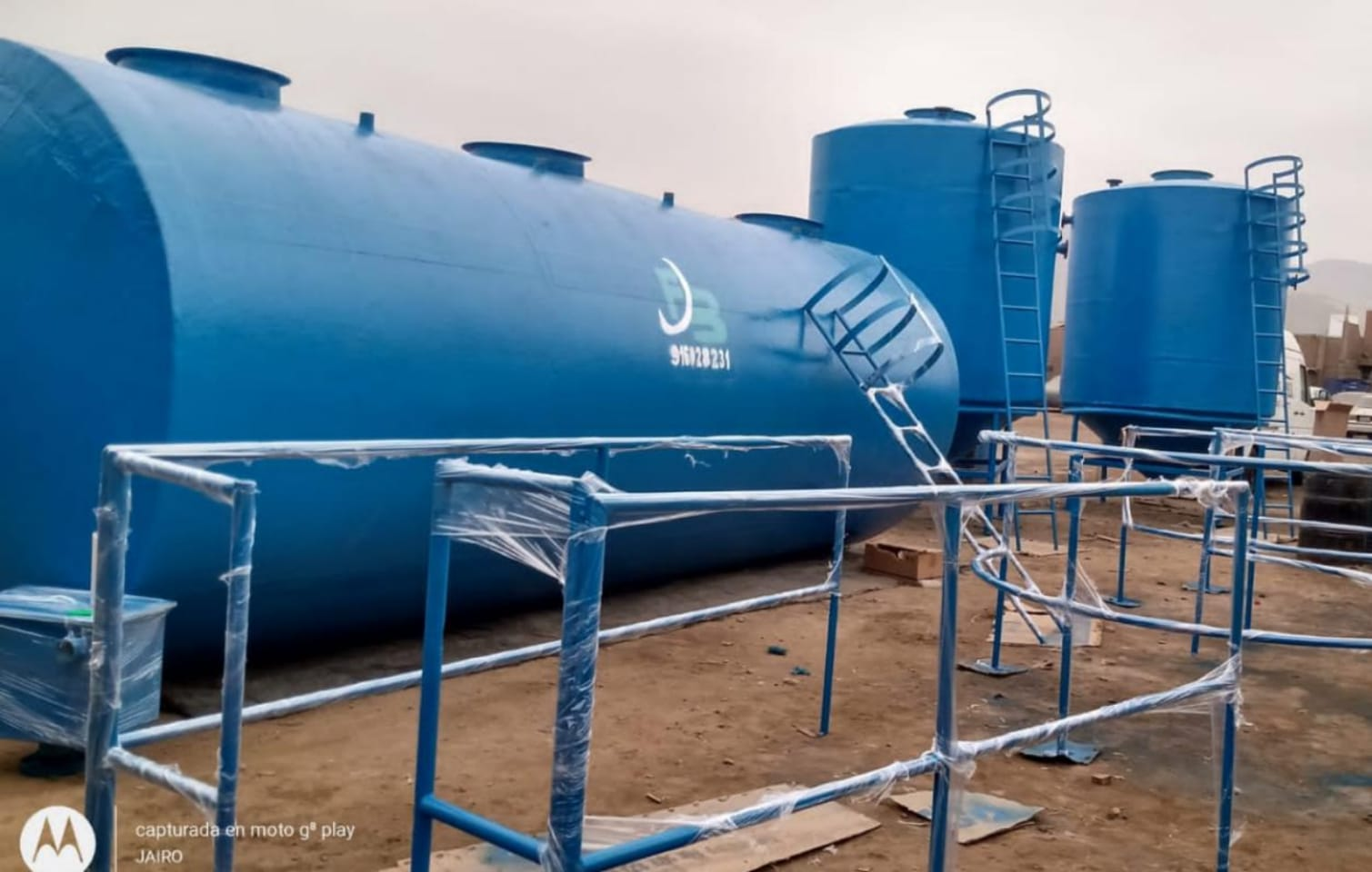 Tanques y tuberías industriales azules en una instalación al aire libre sobre terreno de tierra.