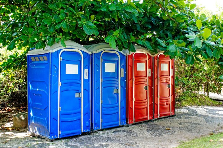 Blue and red portable toilet hire units in outdoor park setting under trees.