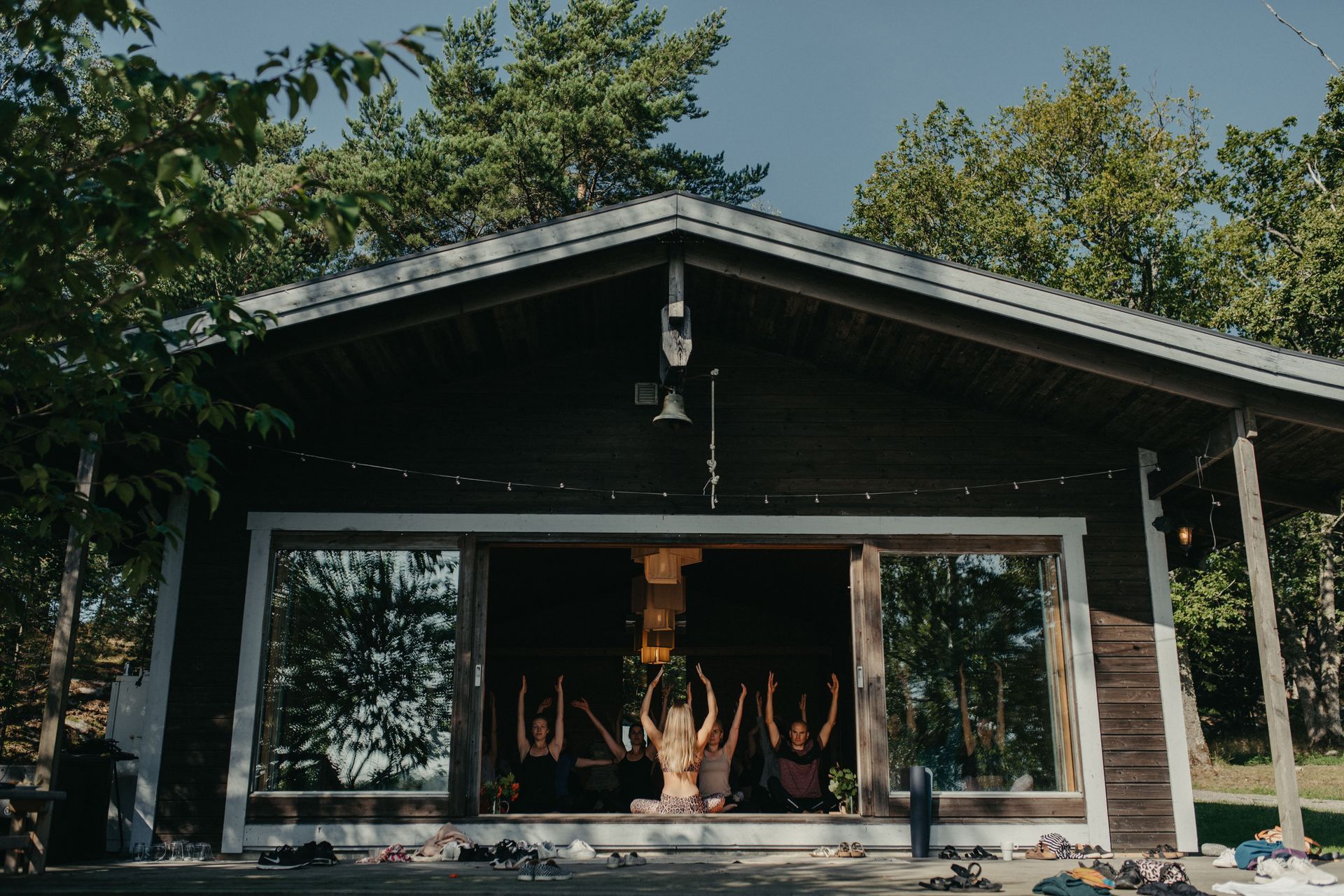A group of people performing yoga inside a wooden studio with open glass walls, surrounded by trees on a sunny day.