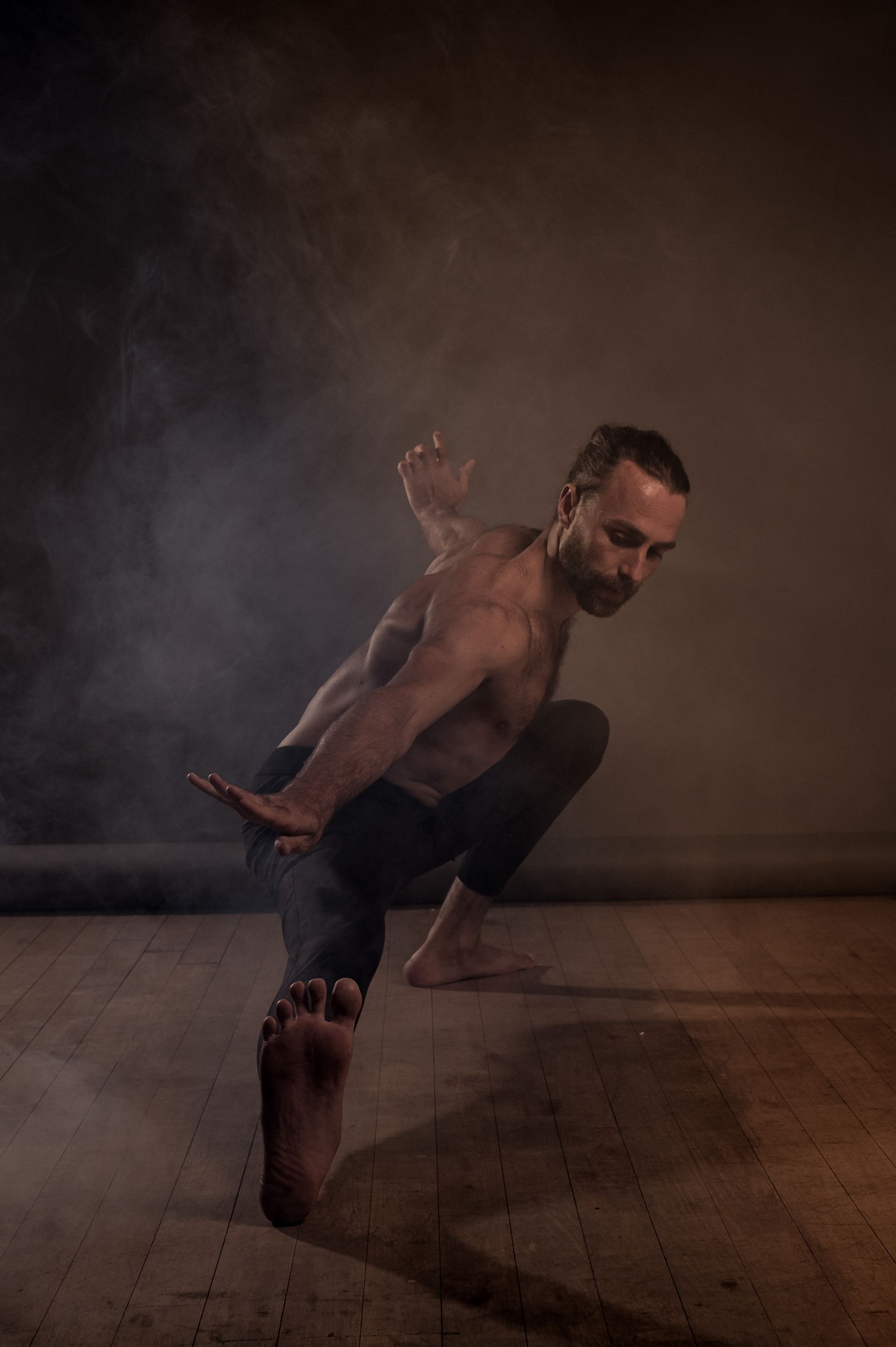 Man in yoga pose, lifting his body. Bare chest, dark pants. Smoke fills the dim-lit space.