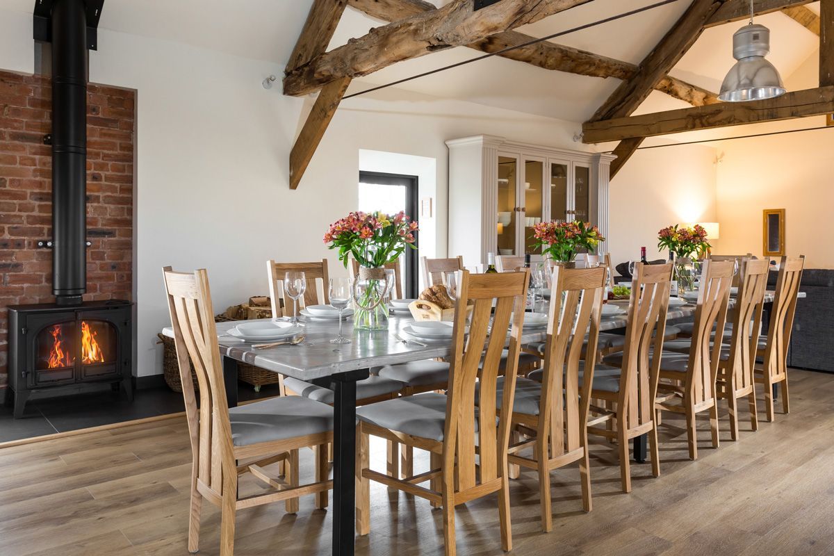 Dining room with long table set for a meal, wooden chairs, fireplace, exposed beams, and flowers.