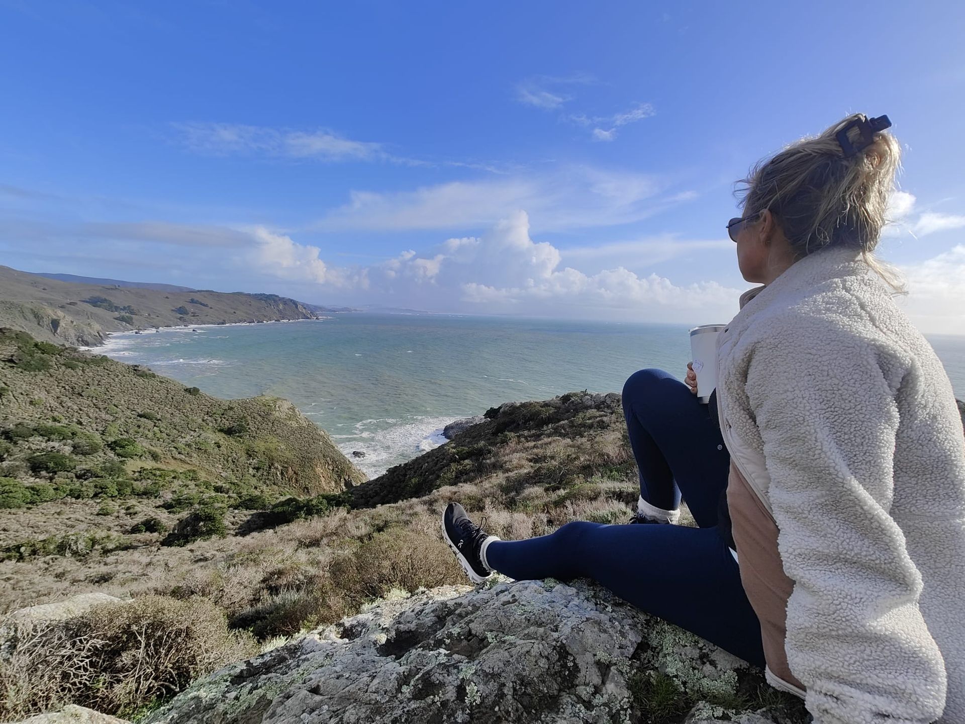 Woman sitting on a cliff, looking at the ocean under a bright blue sky.
