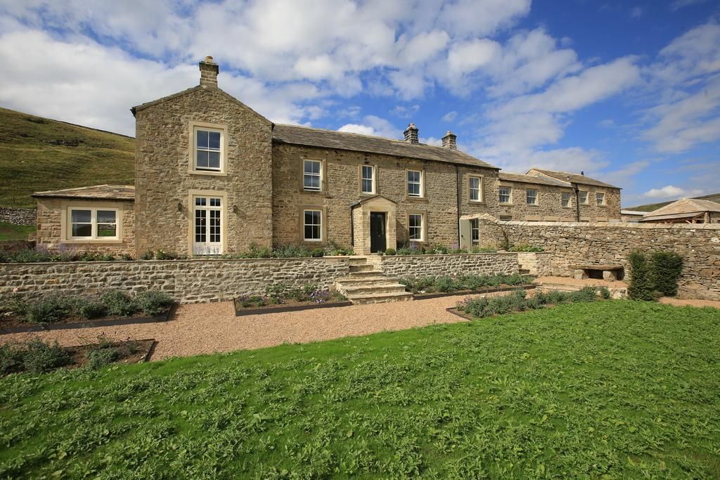 Stone farmhouse on a sunny day with green lawn, flower beds, and a blue sky.