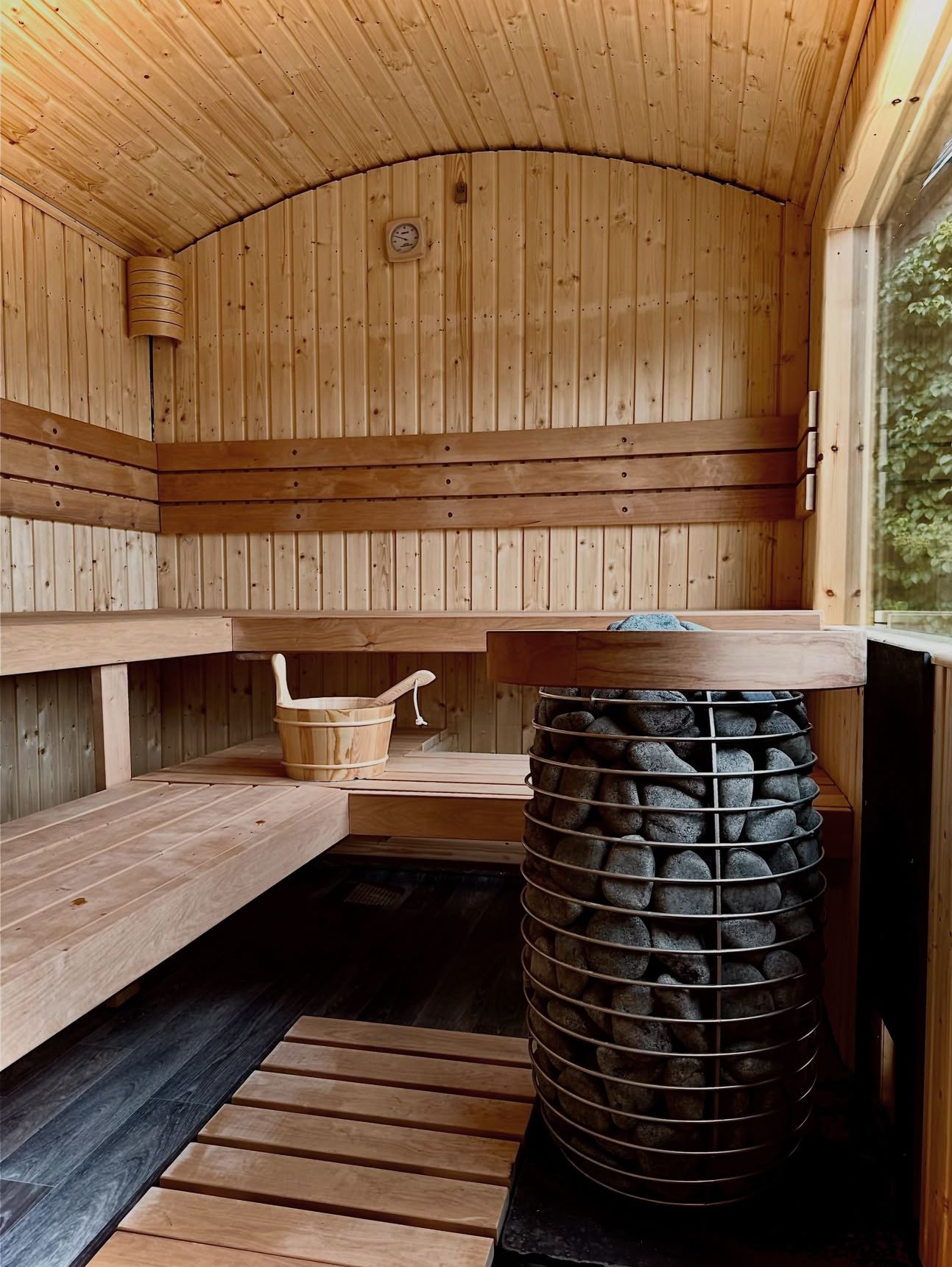 A modern, glass-walled cabin sits on a wooden deck surrounded by trees, connected by a path over rocky terrain.