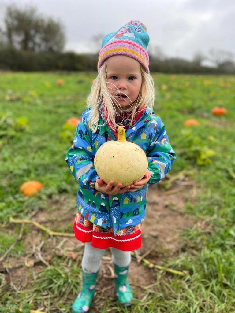 Child holding a light-colored pumpkin in a pumpkin patch, wearing a blue jacket, red dress, and green boots.