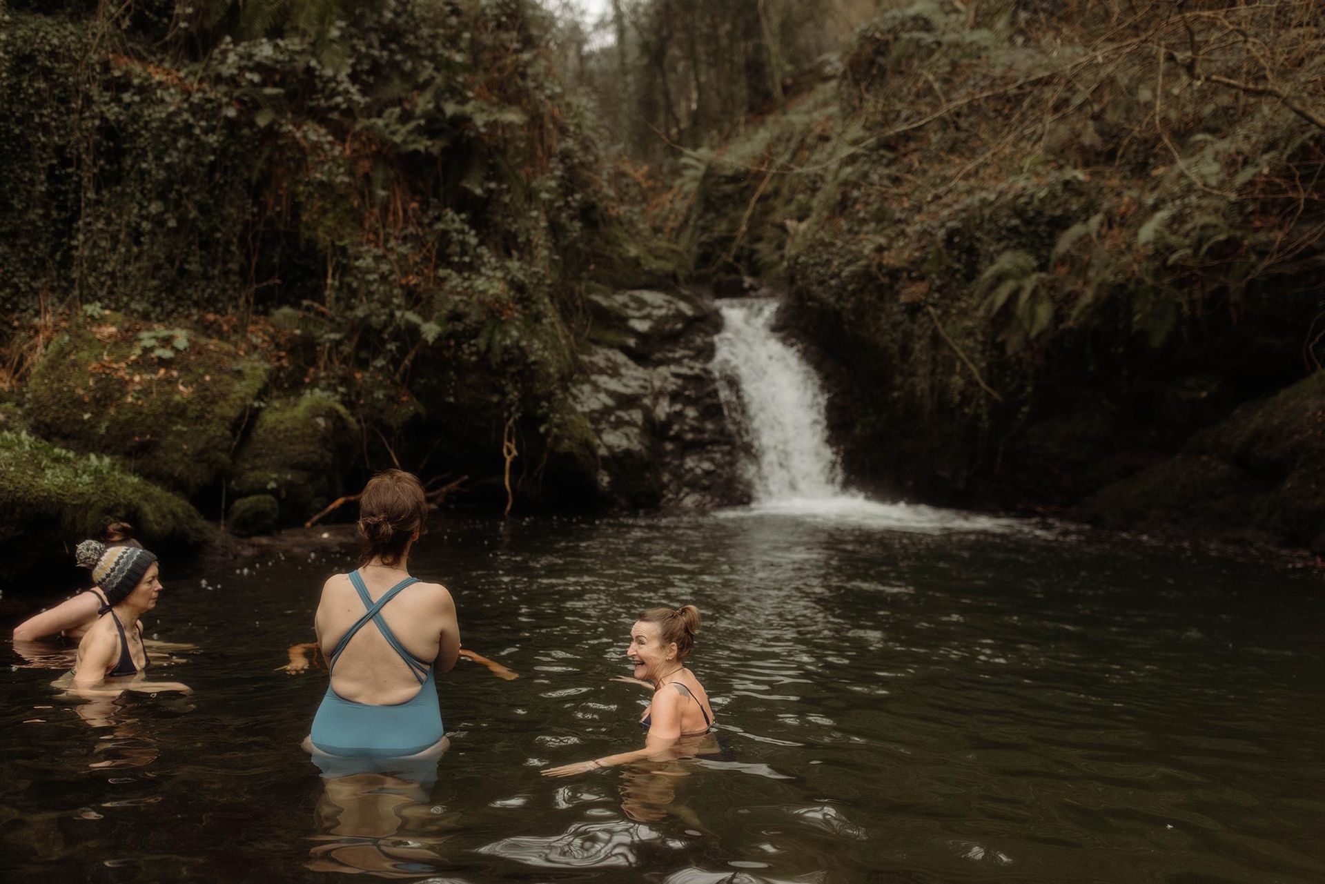 Three people swim in a pond under a small waterfall, surrounded by green trees and rocks.