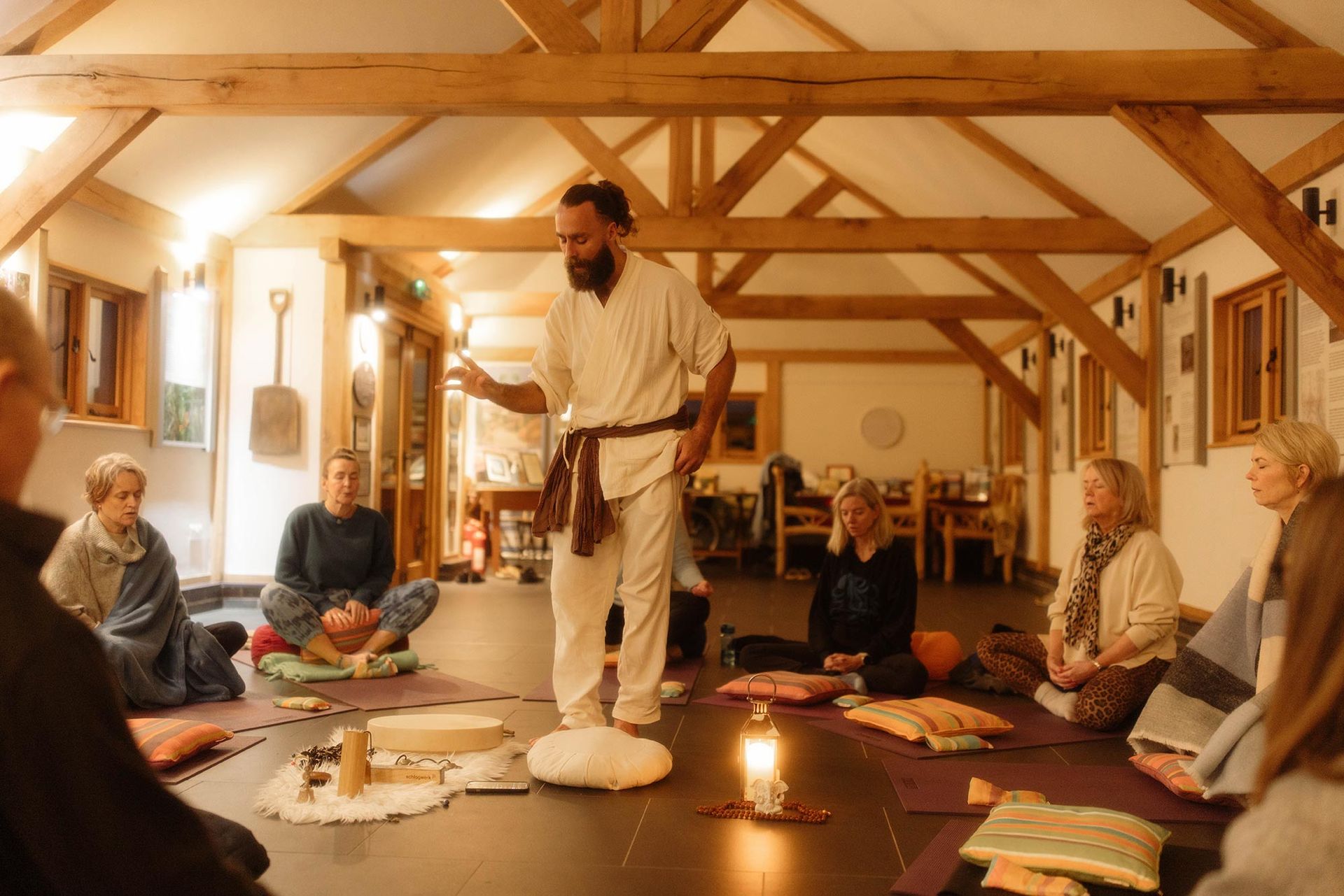 A man in white speaks to a seated group inside a wooden-beamed building.