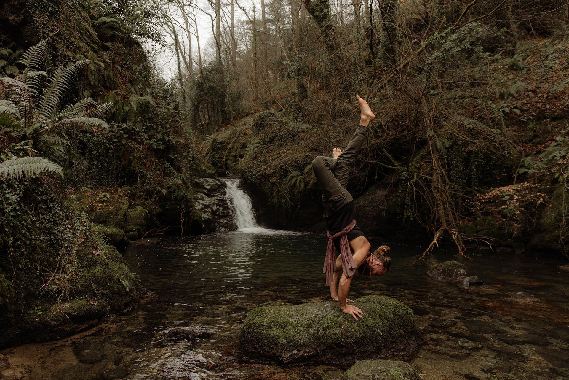 Person doing a handstand on a mossy rock in a forest stream near a small waterfall.