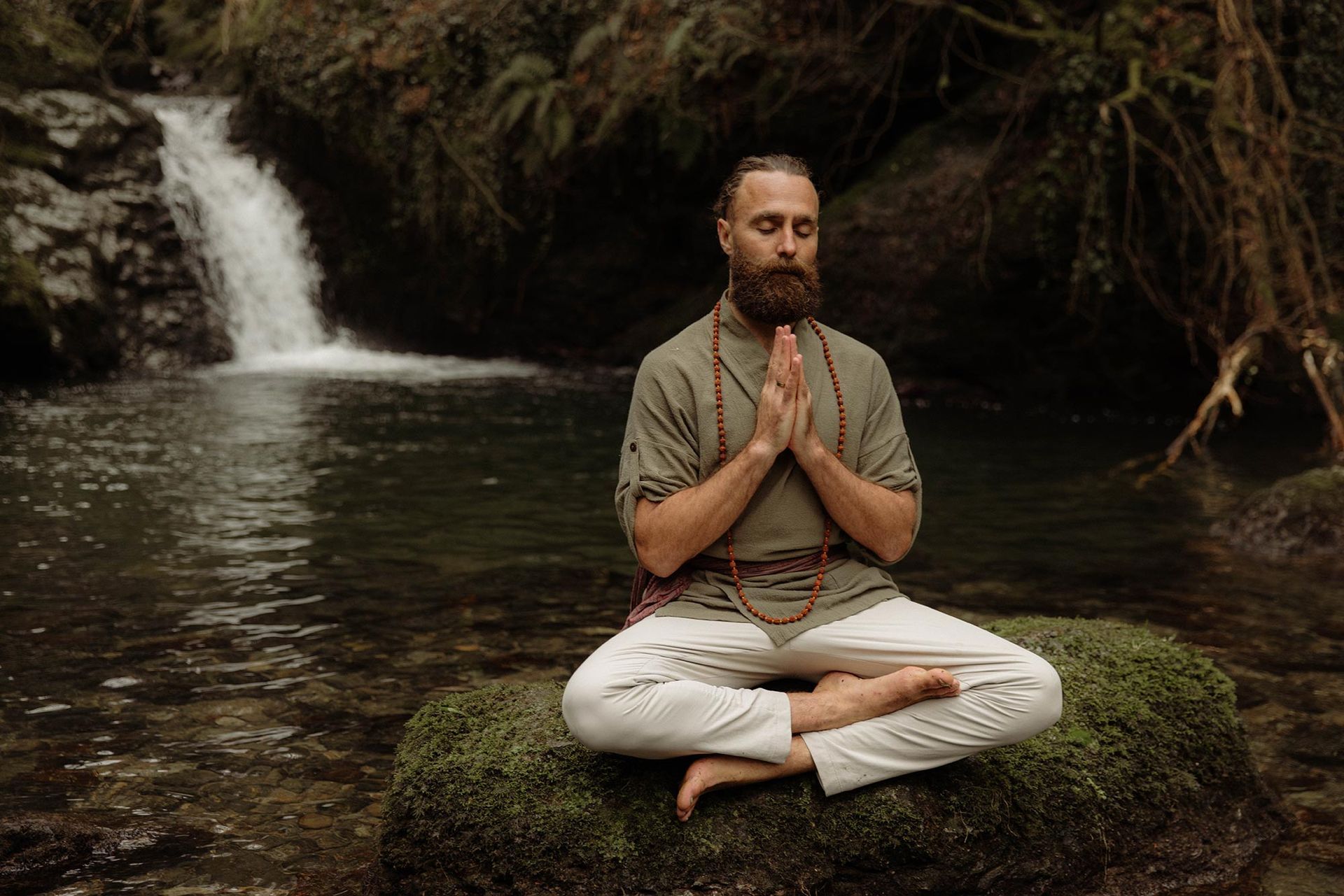 Man meditates with hands together near waterfall. He's wearing a green shirt, white pants, and a beaded necklace.