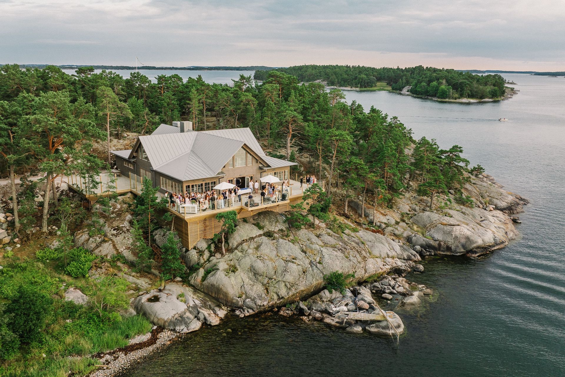 Aerial view of a coastal event venue with a grey roof situated on a rocky island surrounded by water and pine trees.