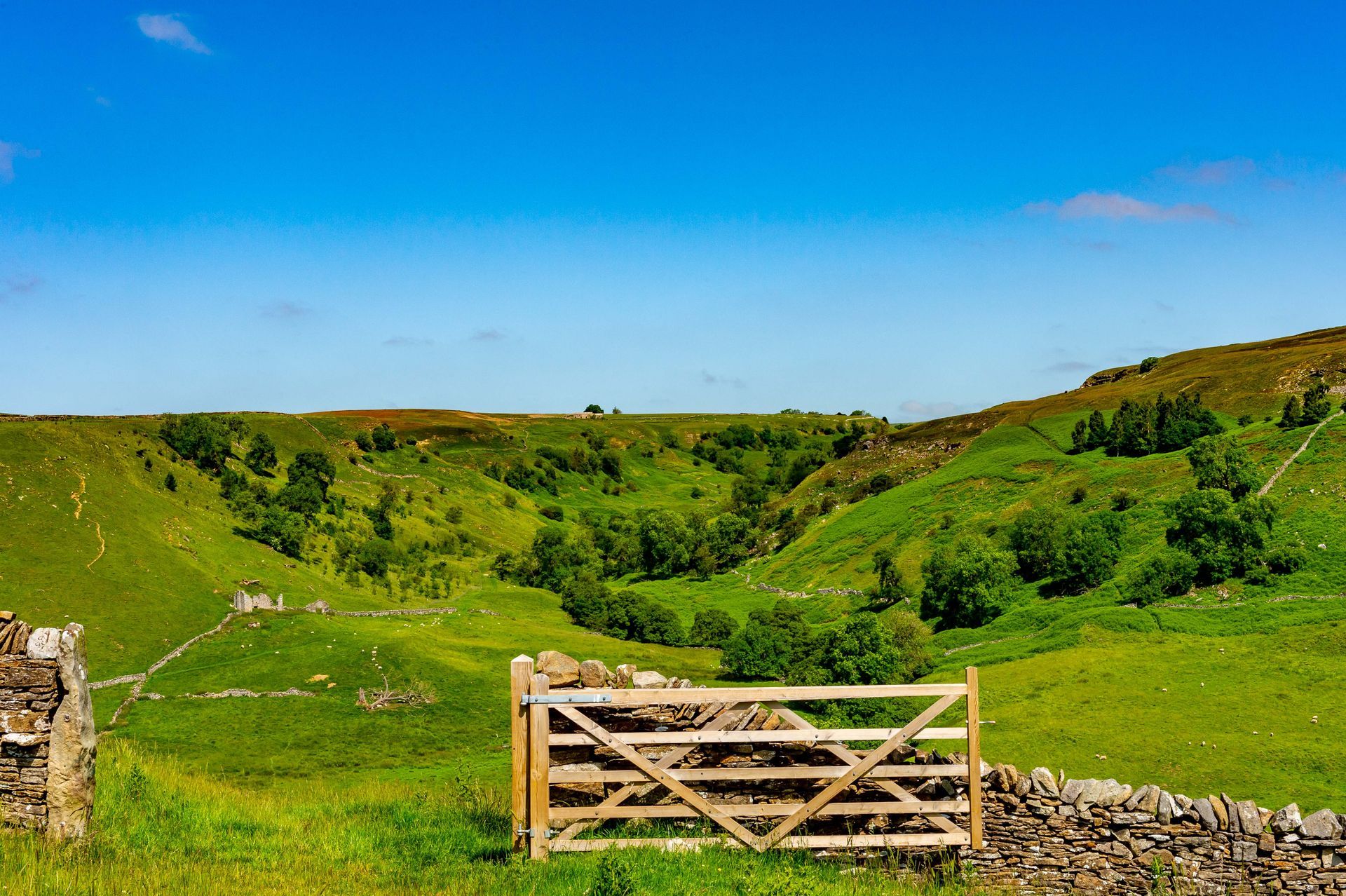 Rolling green hills with a wooden gate, stone walls, and a blue sky.