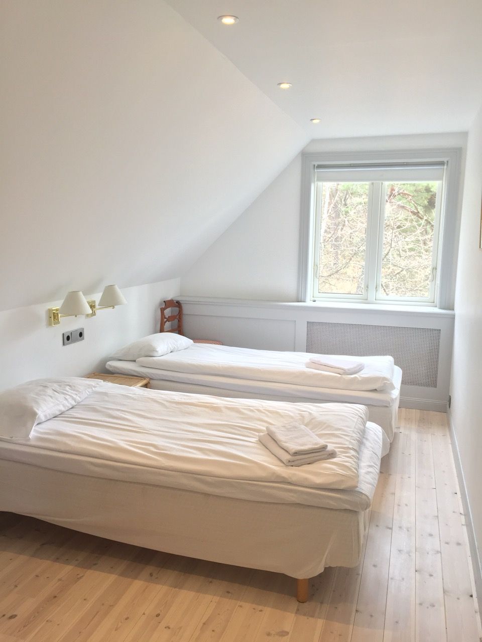 A bright, minimalist bedroom with two single beds, white bedding, light wood floors, and a window with a sloped ceiling.