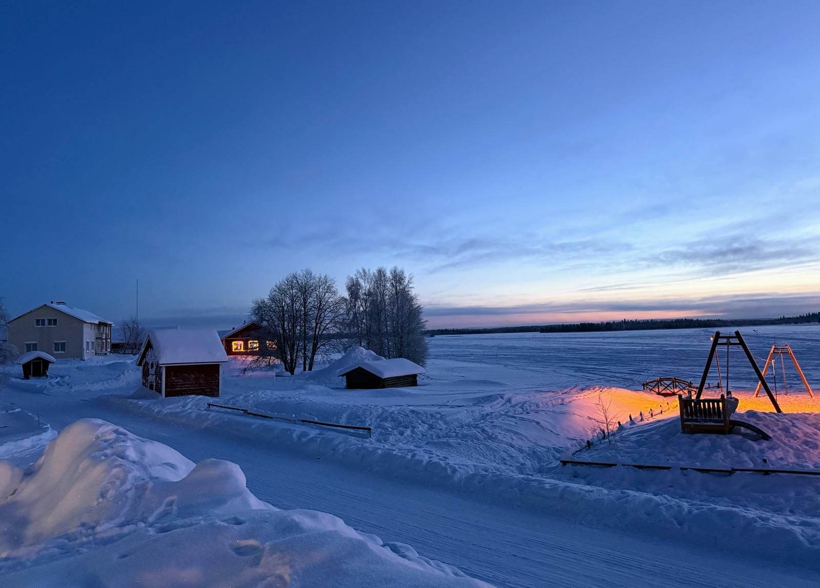Snowy landscape with buildings and a frozen lake under a twilight sky.