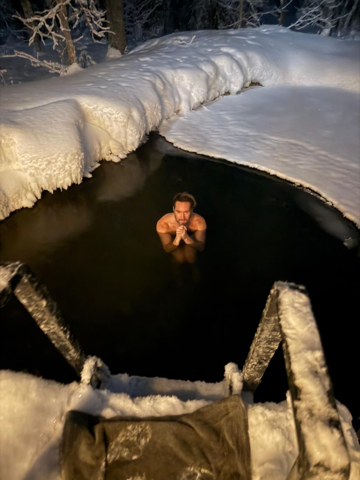 Man in dark water, surrounded by snow-covered banks, climbs out with towel on wooden steps.