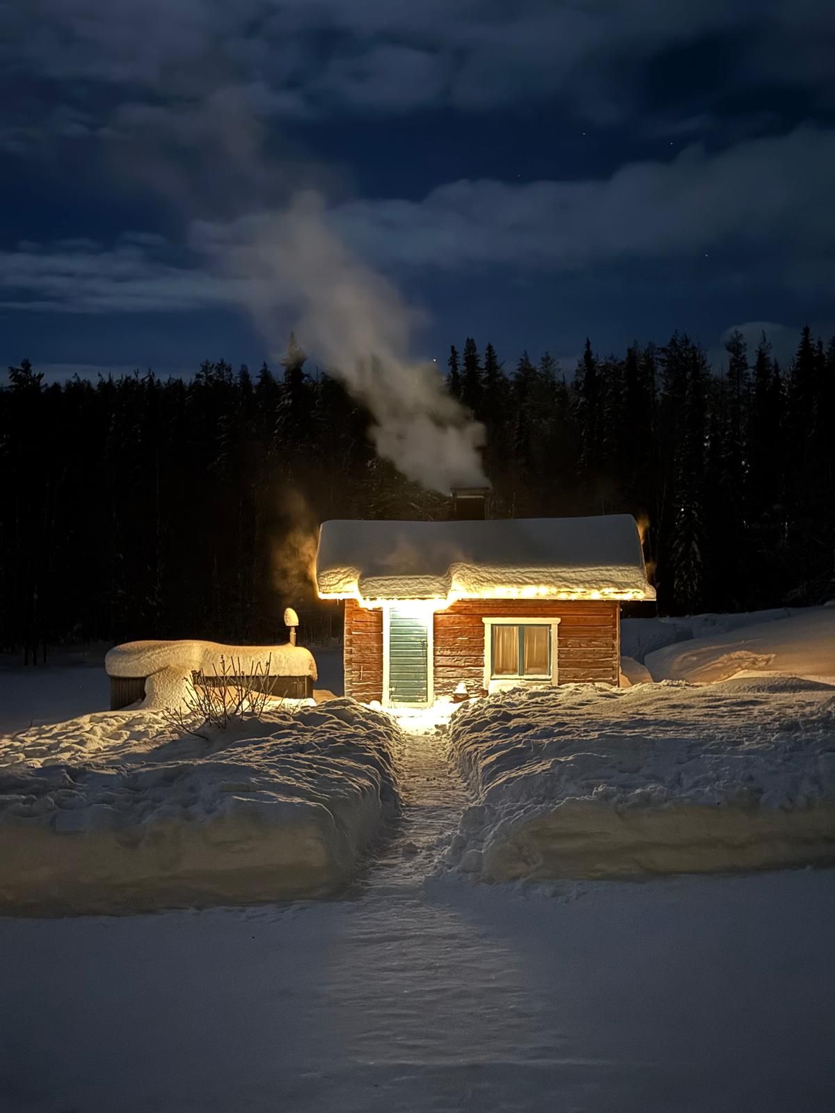 Red cabin in snow with lit doorway and smoking chimney at night.