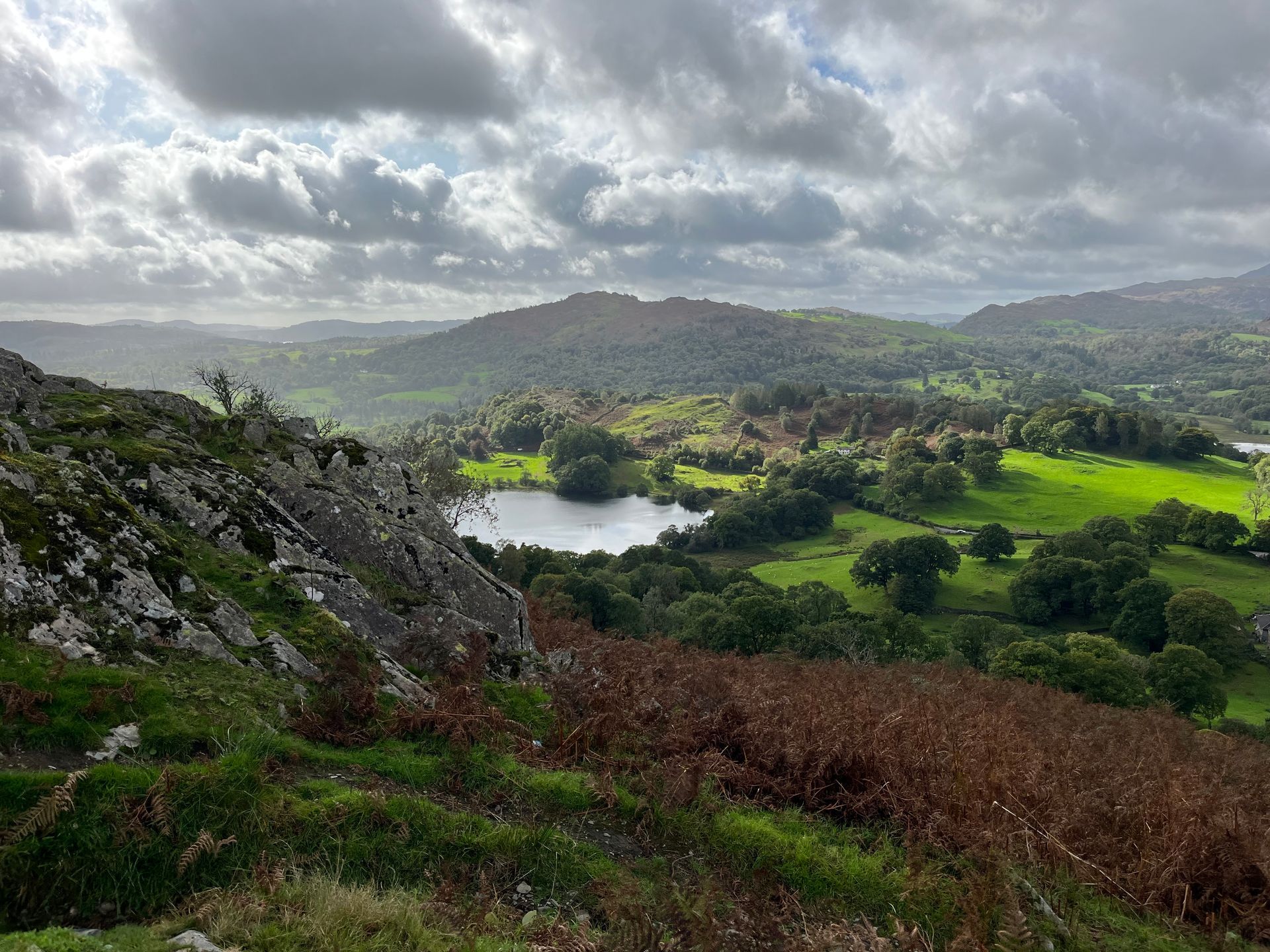 A small lake set in a lush green valley, viewed from a rocky, fern-covered hillside under a dramatic cloudy sky.