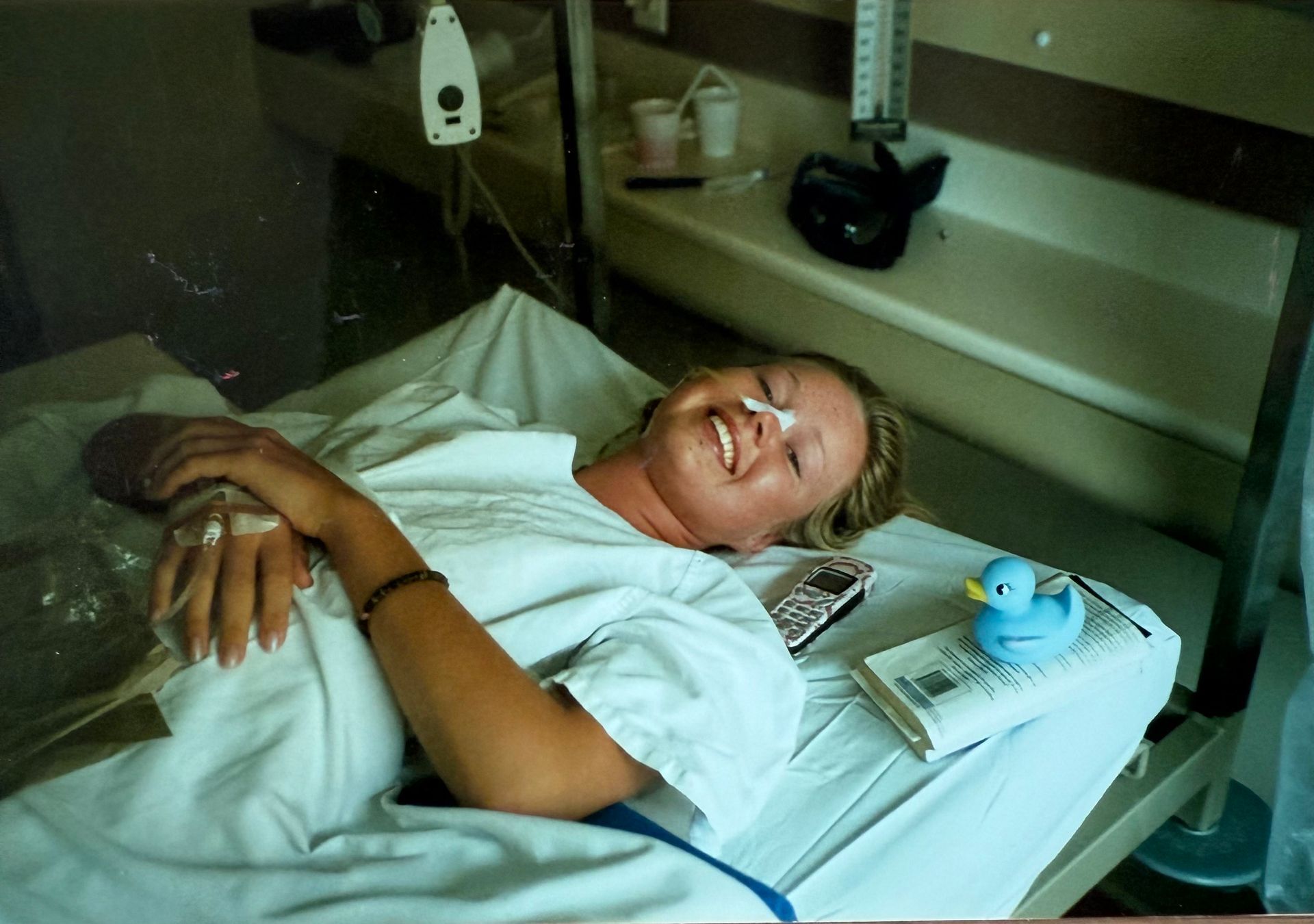 Woman in hospital bed smiling, with IV, oxygen tube, and a rubber duck.