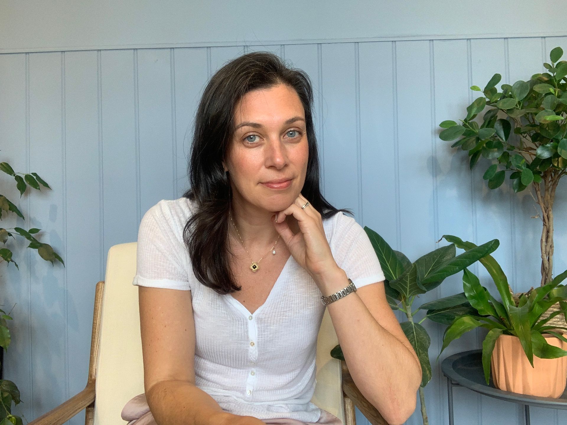 Woman in white shirt sitting, hand on neck, smiling; plants and blue wall in background.