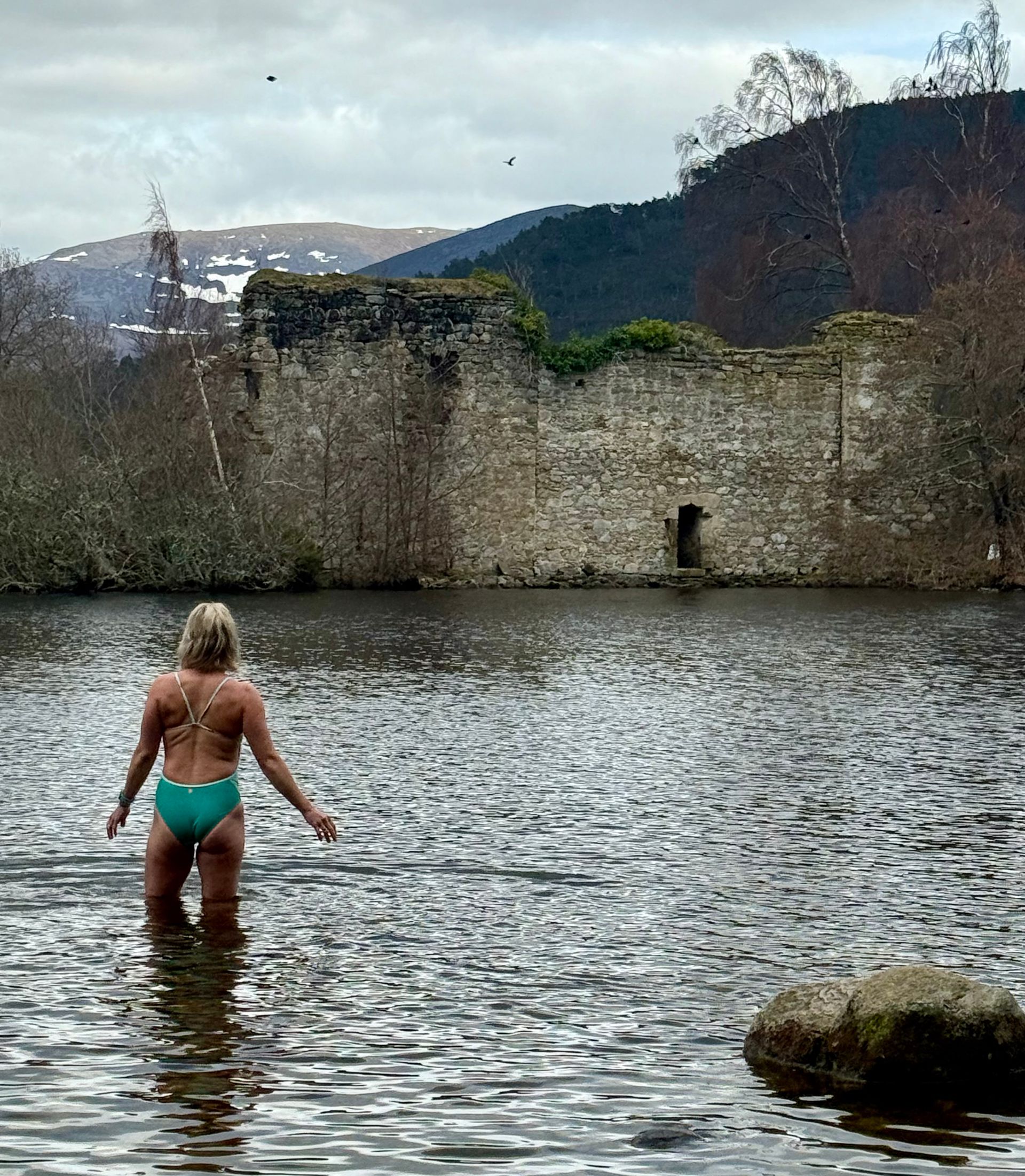 A person in a turquoise swimsuit stands in a lake facing the stone ruins of a castle set against hills and cloudy sky.