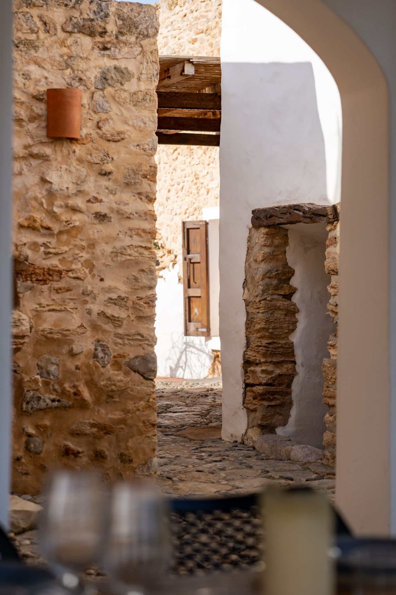 Stone alleyway framed by an arch, leading to a shuttered window, viewed from a blurry table setting.