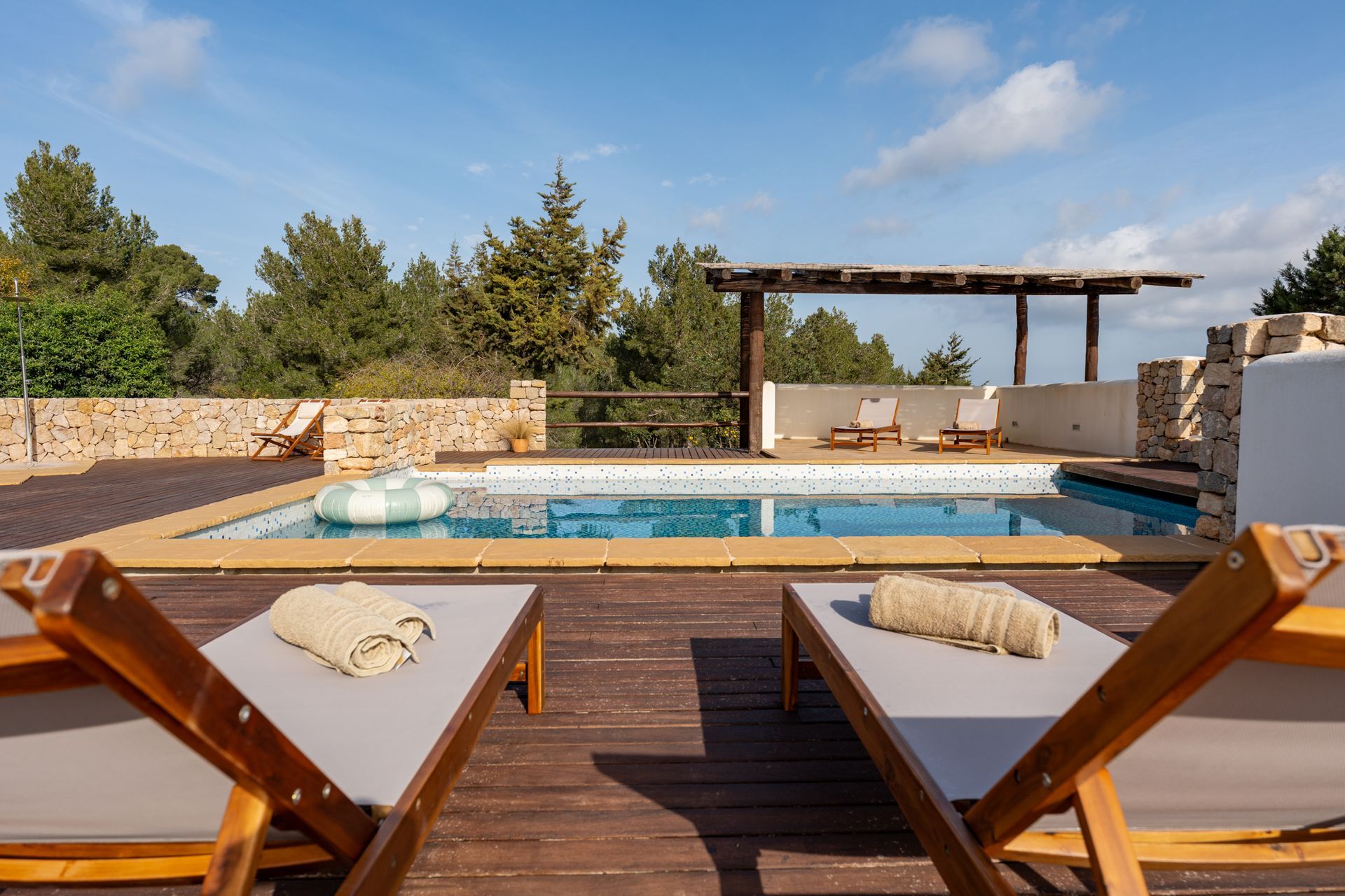 Two lounge chairs on a wooden deck overlook a pool with a pergola and trees in the background.