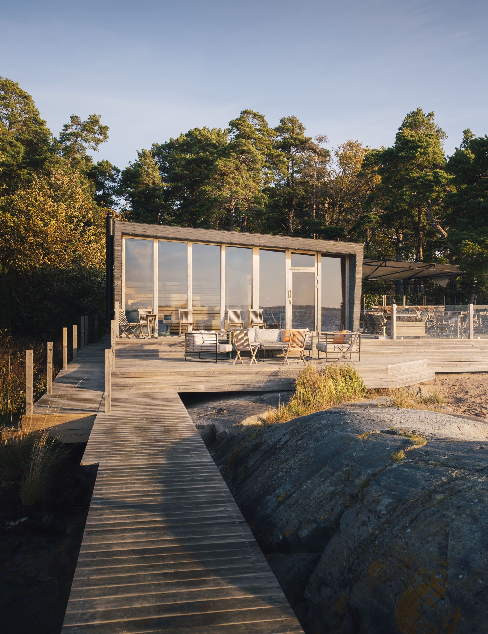 A modern, glass-walled cabin sits on a wooden deck surrounded by trees, connected by a path over rocky terrain.