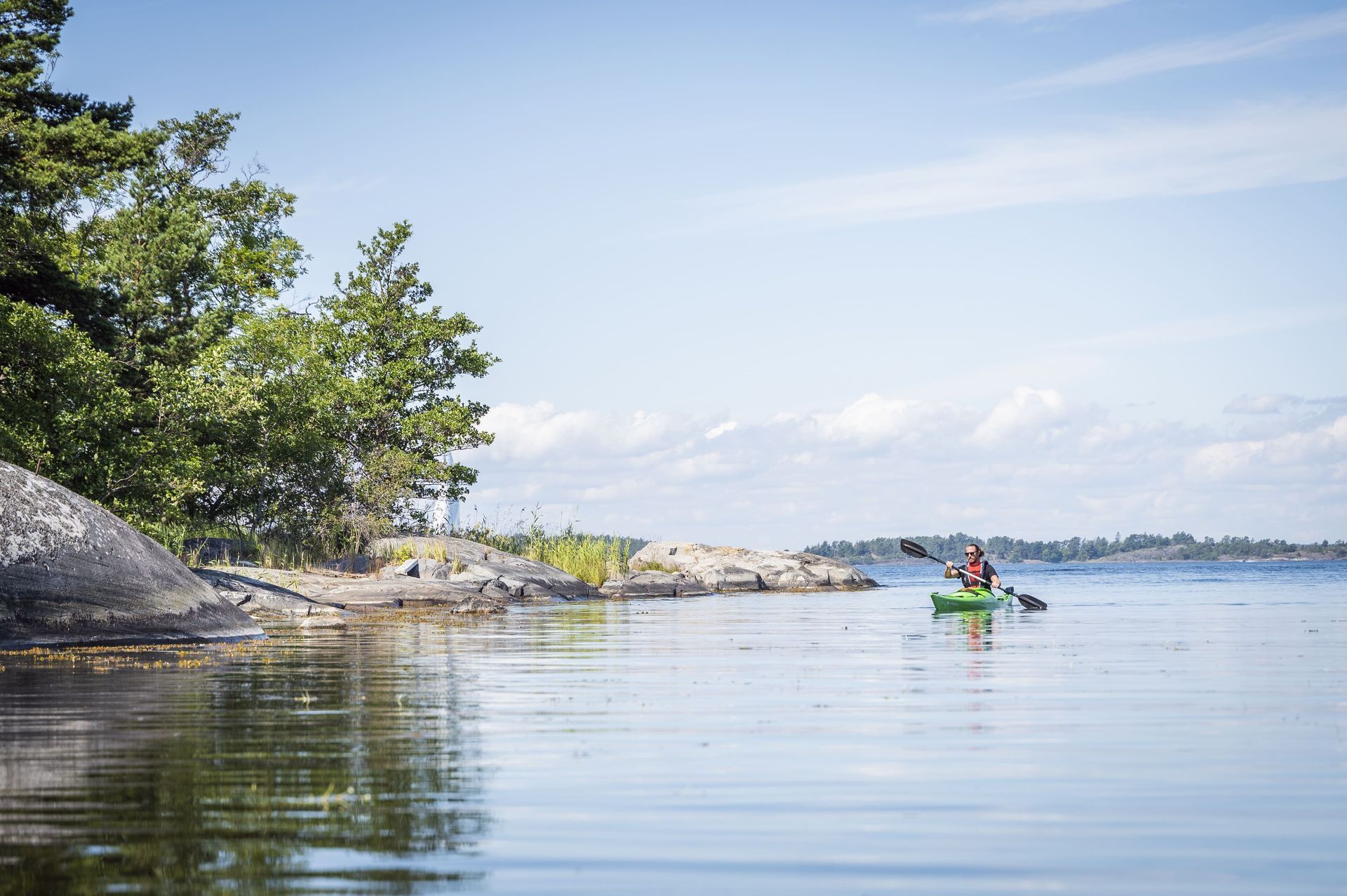 A person paddles a green kayak on calm water past a rocky, tree-lined coastline under a clear blue sky.