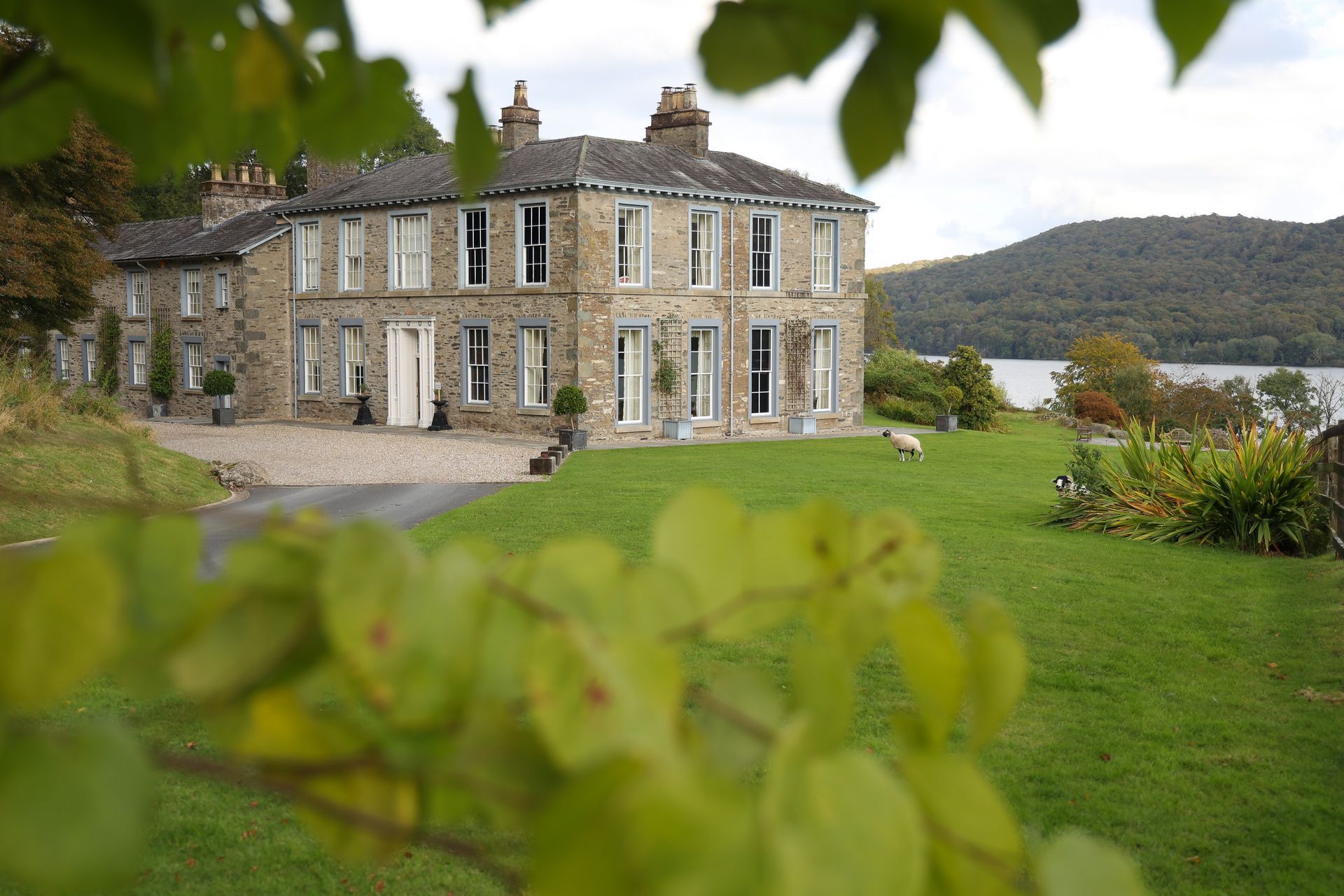 Large stone house with white window frames and a green lawn, overlooking a lake and hills.