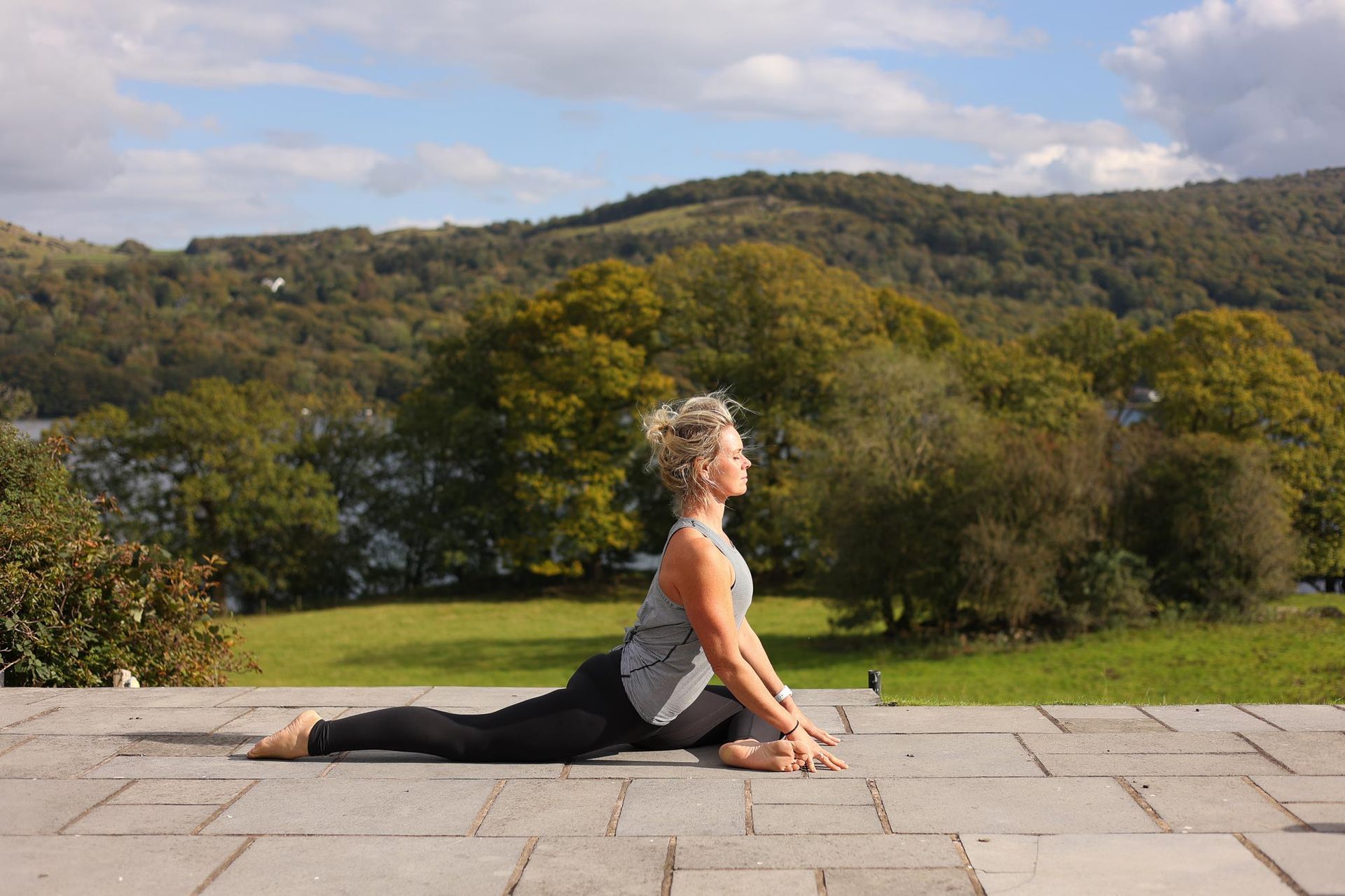 Woman in yoga pose, seated with one leg extended, outdoors with trees and a mountain in the background.