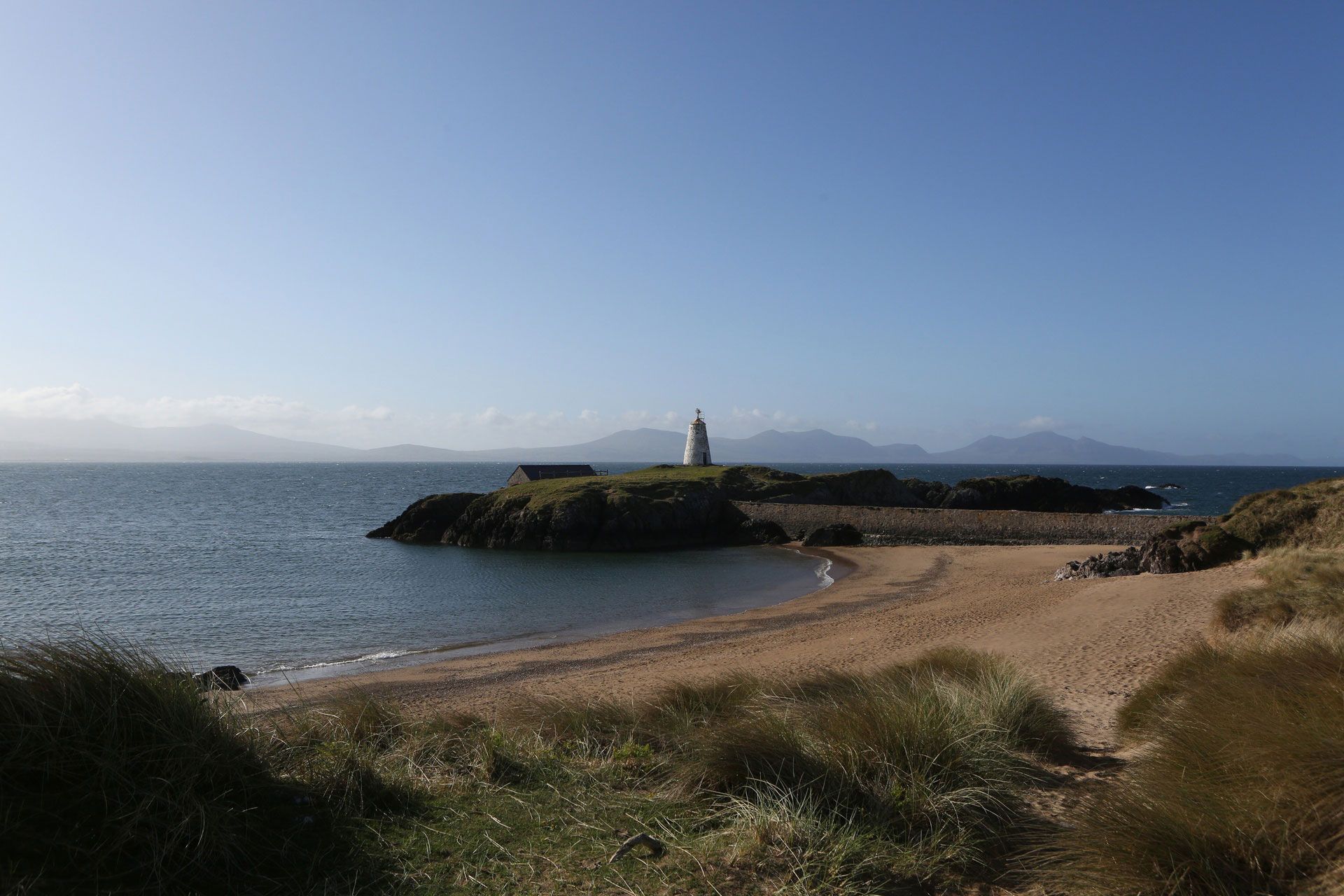 Seascape with a sandy beach, rocky island with a lighthouse, and mountains under a blue sky.