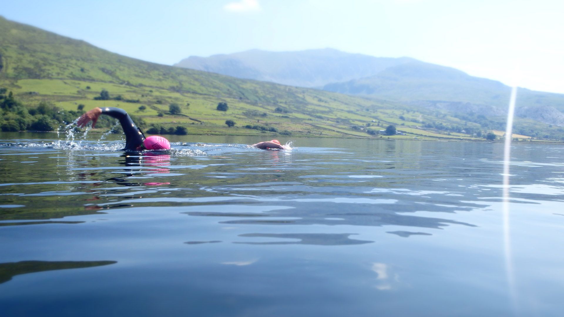 Two swimmers in a lake, one wearing a pink cap and black wetsuit, with a mountain backdrop.