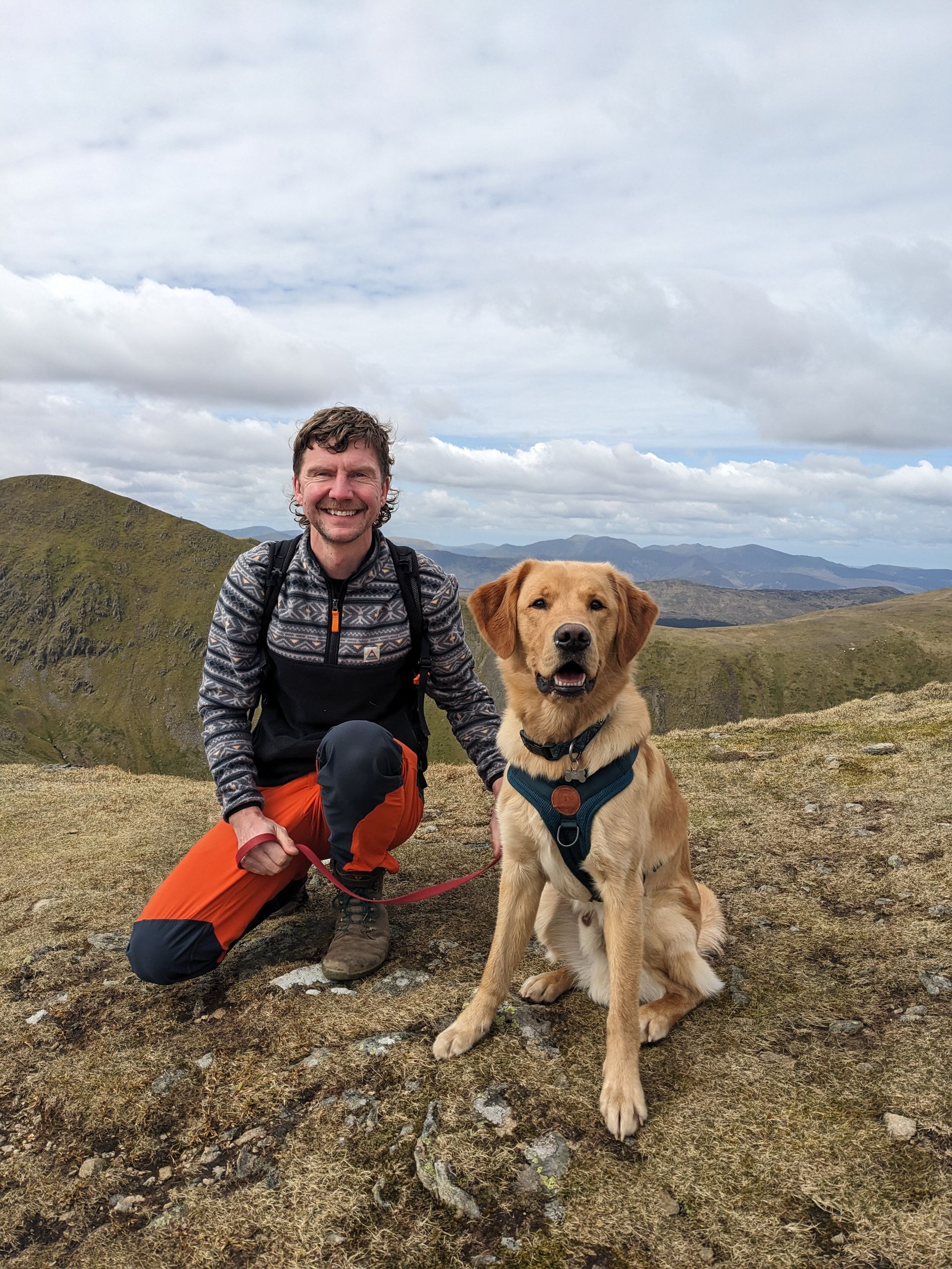 Man kneels next to golden retriever on a mountain top, smiling. Overcast sky, distant mountains.
