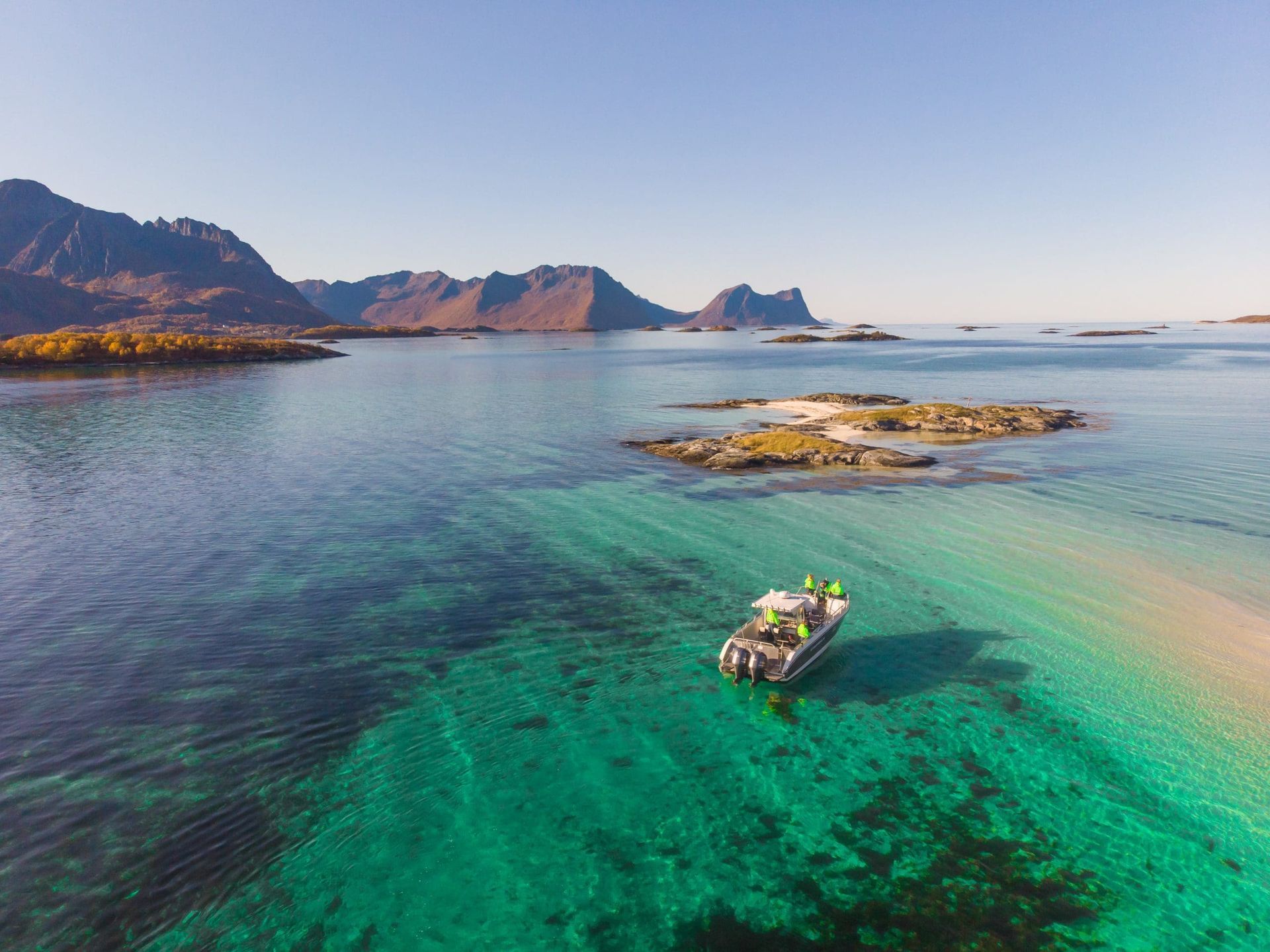 Boat in turquoise water near a sandy islet, with mountains in the background under a clear sky.