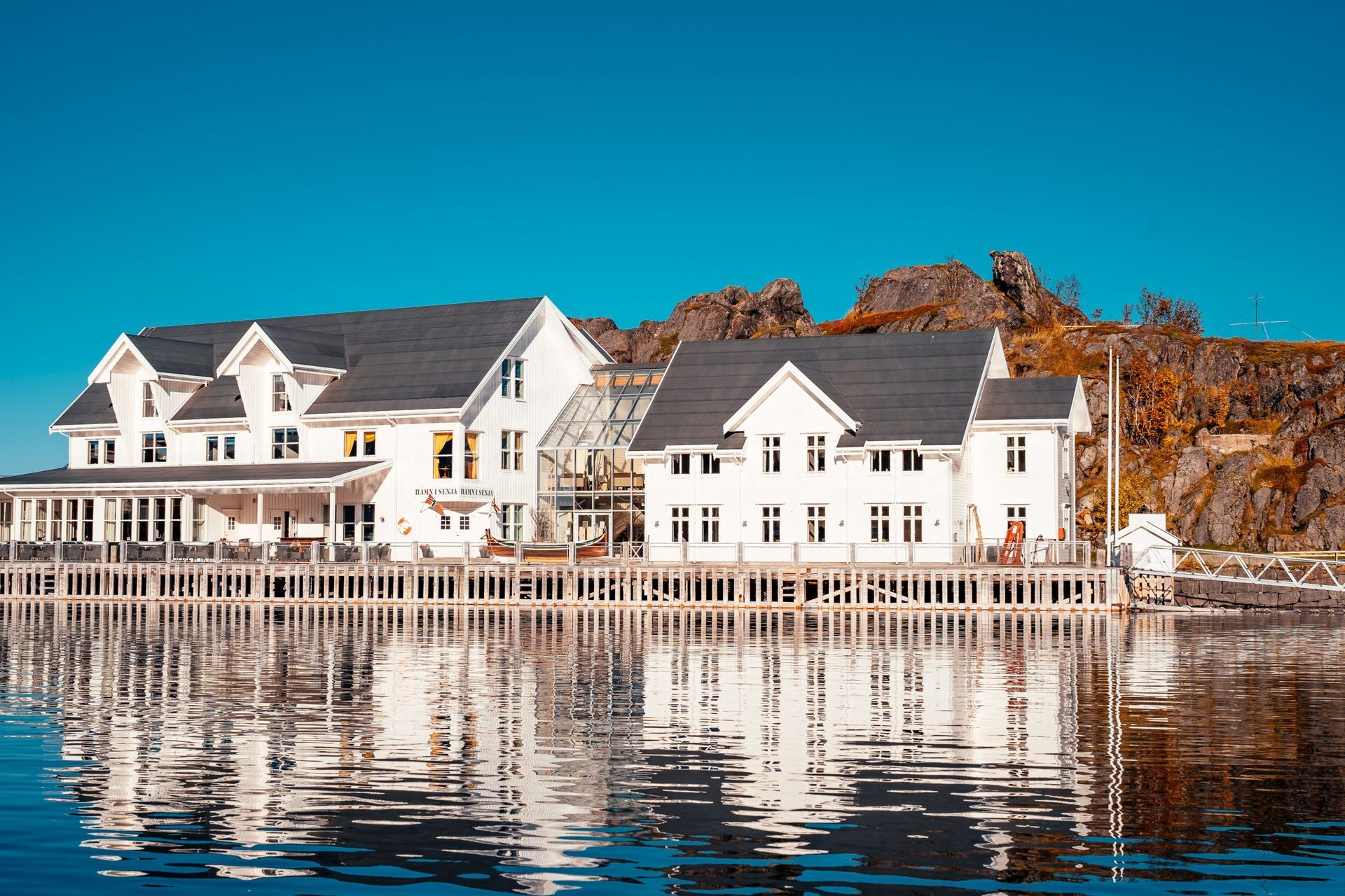 White buildings on a pier reflected in calm water, against a rocky backdrop and blue sky.