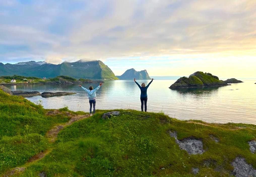 Two people with arms raised stand on a grassy hill overlooking a calm ocean and mountain range.