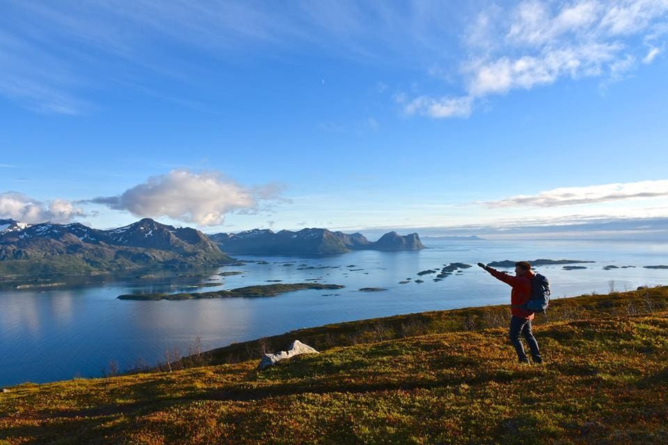 Man with backpack on mountain overlooking a fjord, arms outstretched. Blue sky, water, and mountains.