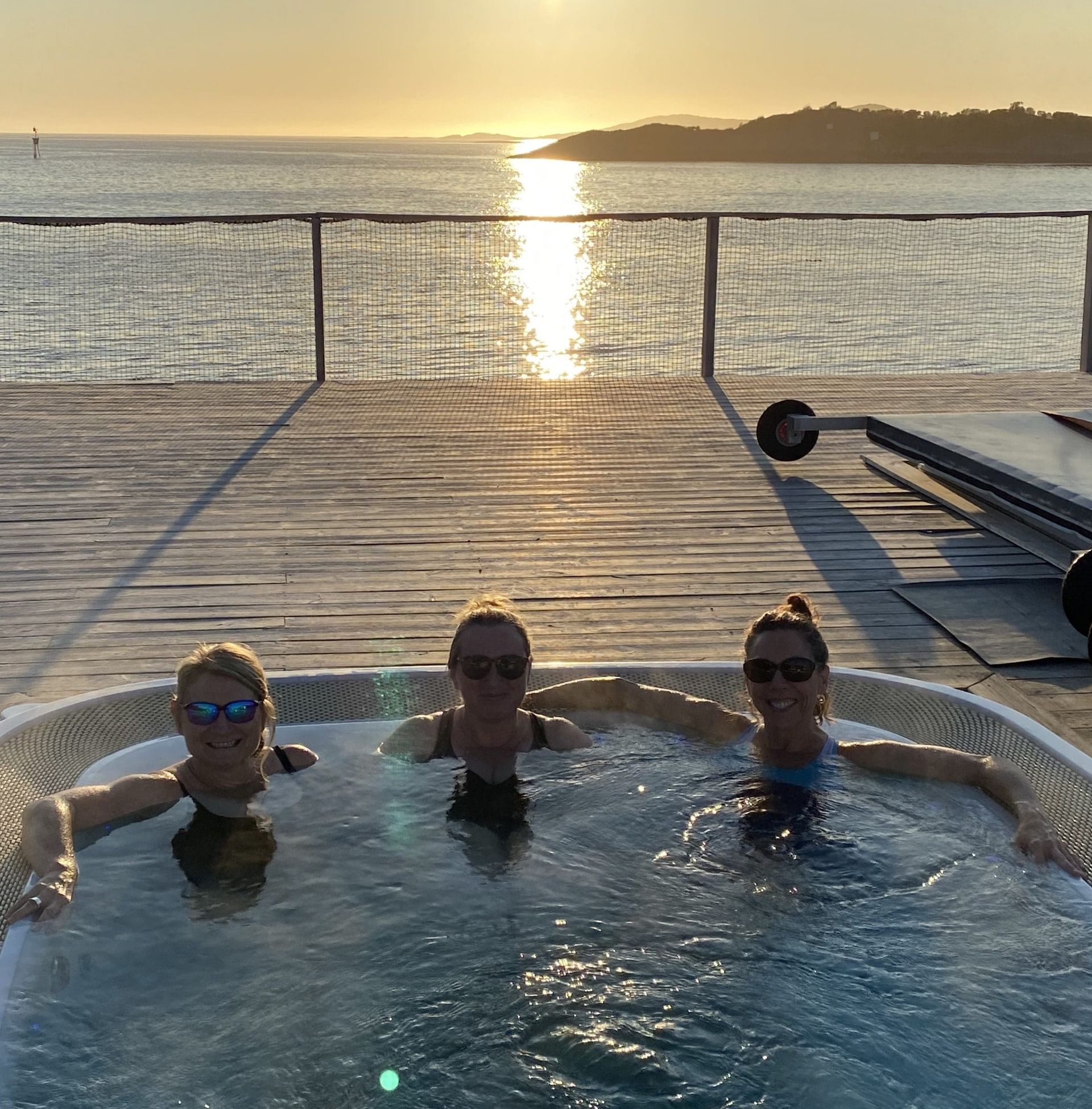 Three people in a hot tub on a deck overlooking the ocean at sunset.