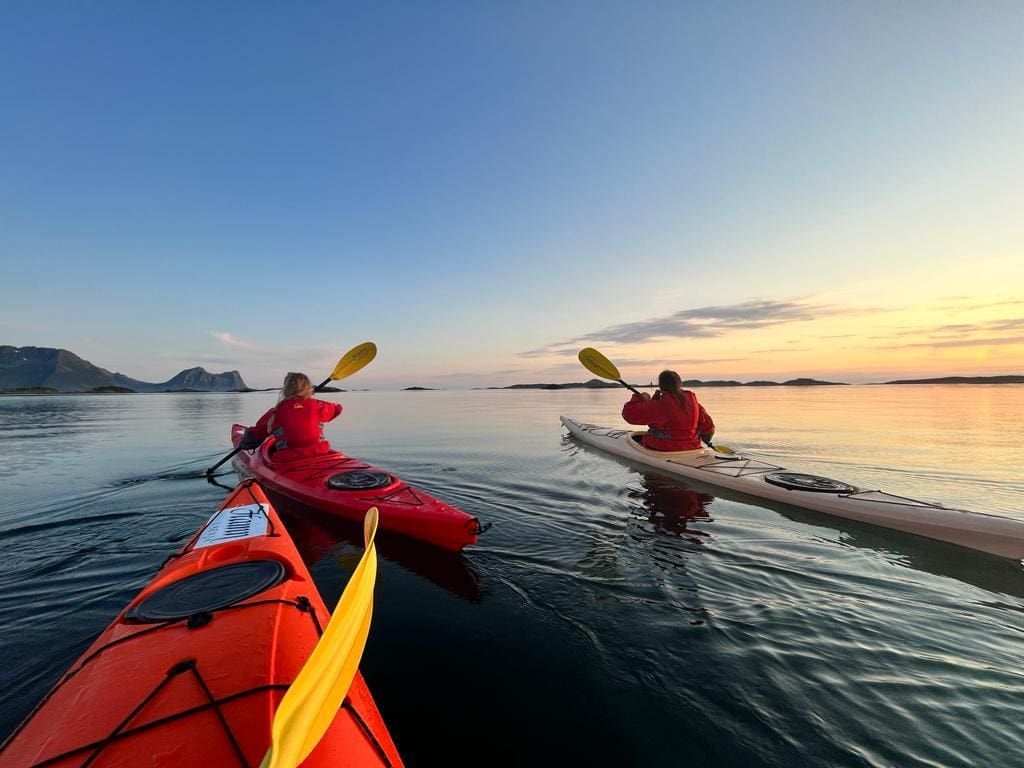 Kayakers on calm water near mountains at sunset, paddling with yellow and green paddles.