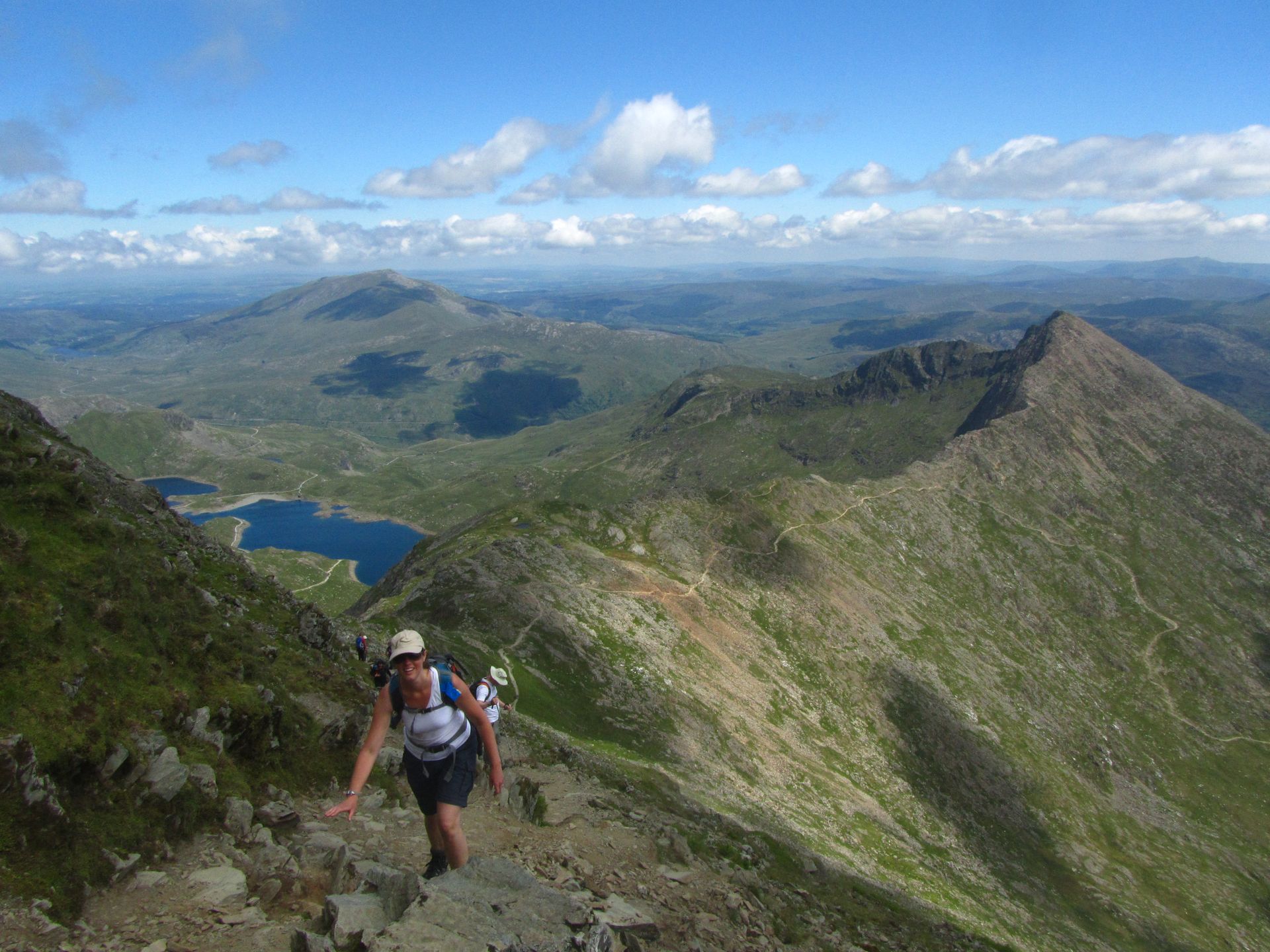 A hiker climbs a steep, rocky trail on a mountain ridge with a large, sweeping valley and blue lakes in the background.