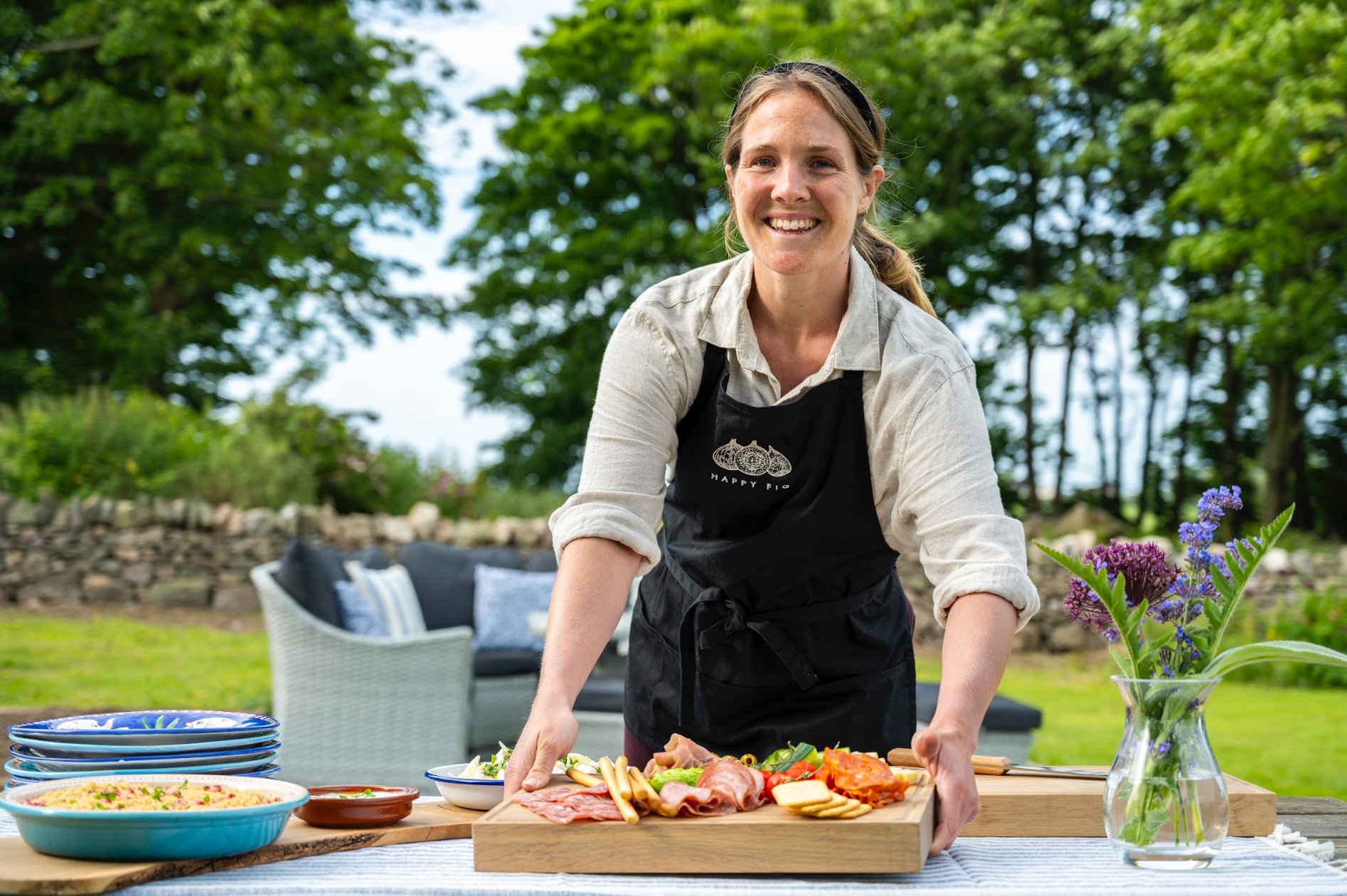 Woman arranging food on a table outdoors; sunny day, smiles.