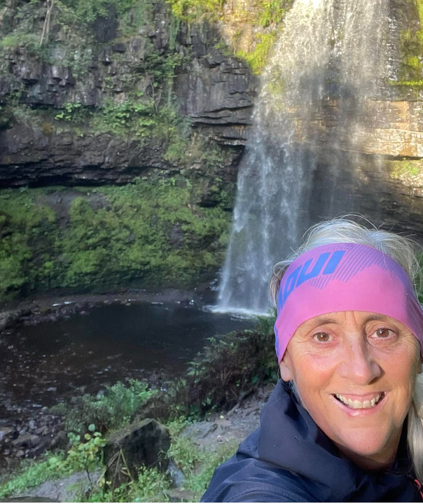 Woman smiles at the camera with waterfall in the background. She wears a pink headband.