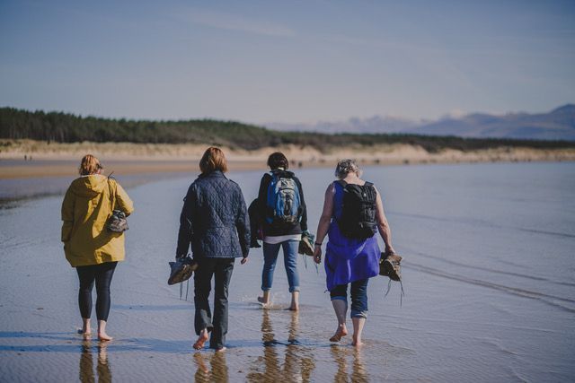 Four people walking barefoot on a wet beach, holding shoes. Mountains and a treeline in the distance.