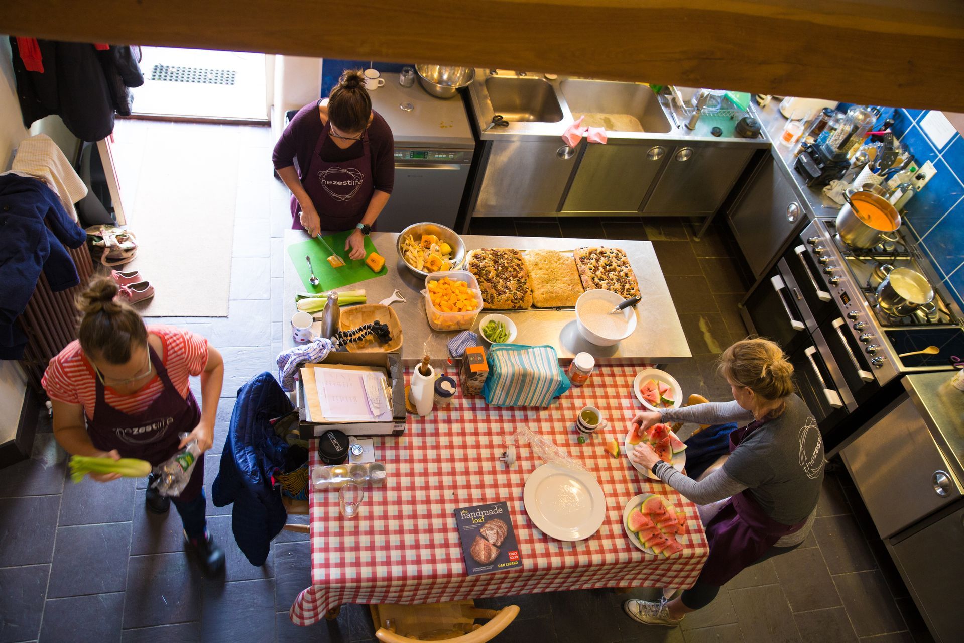 A high-angle view of three people preparing food in a kitchen with a checkered tablecloth, metal counters, and a stove.