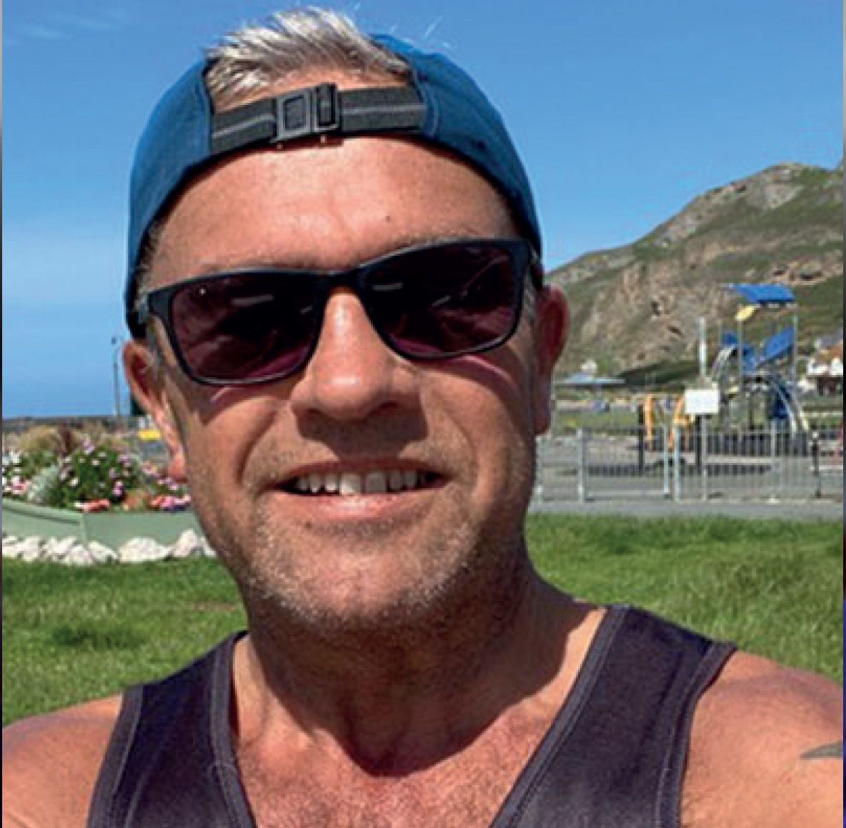 Man in sunglasses and hat smiles outdoors near a park and ocean.