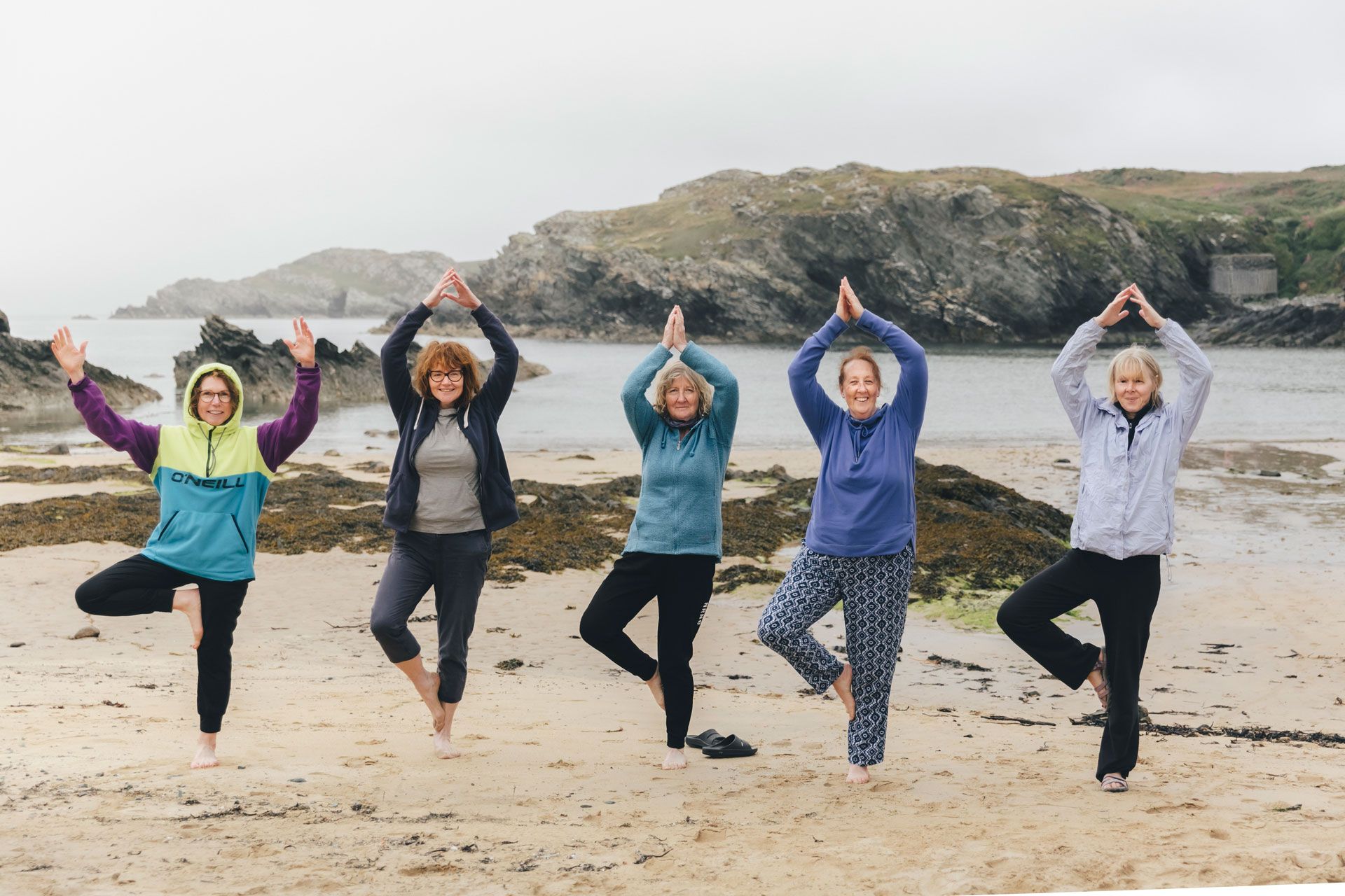 Five people in yoga poses on a beach, arms raised above heads. Cloudy sky, rocky coastline in background.