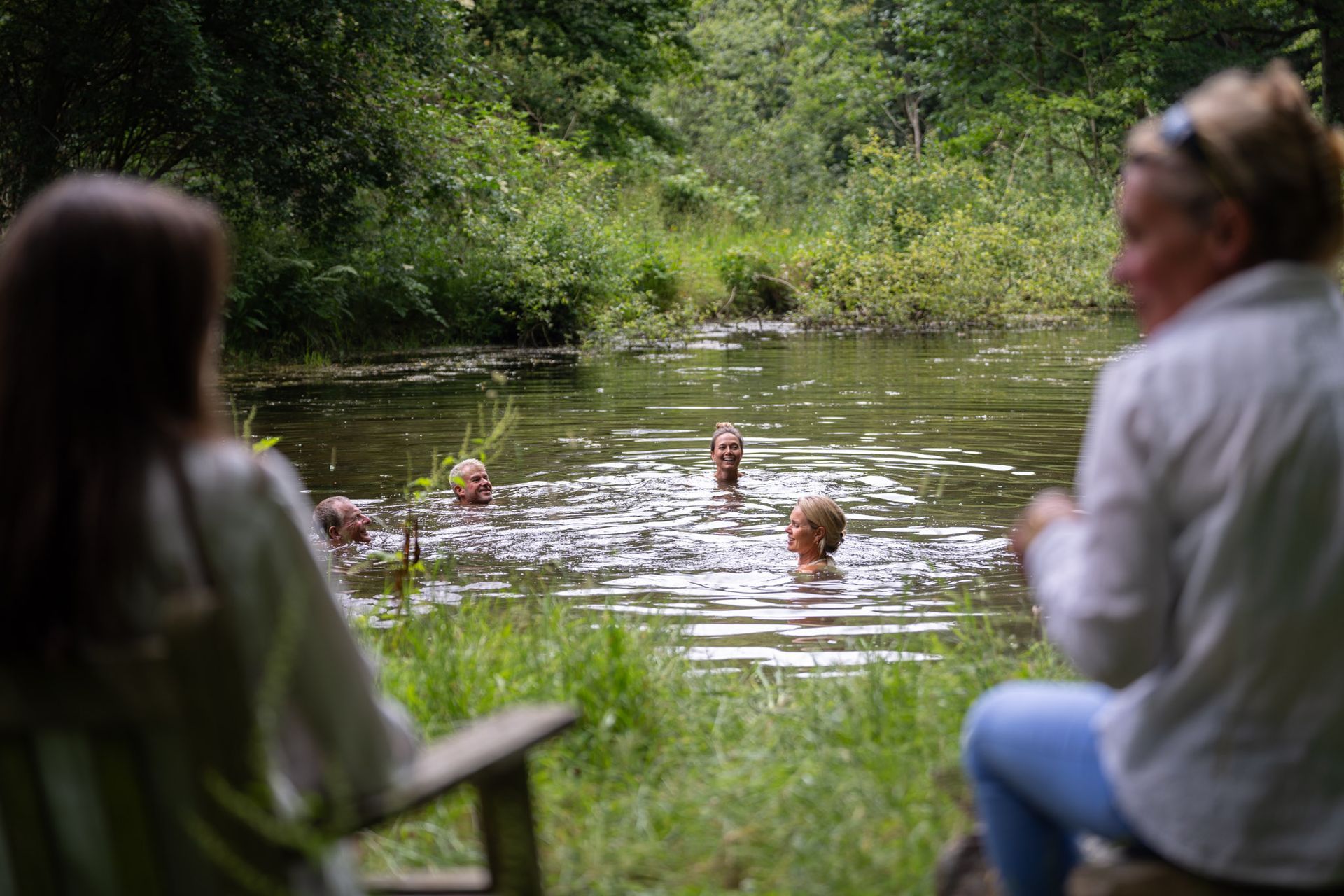People swim in a pond as two others watch from the grassy bank, surrounded by trees.