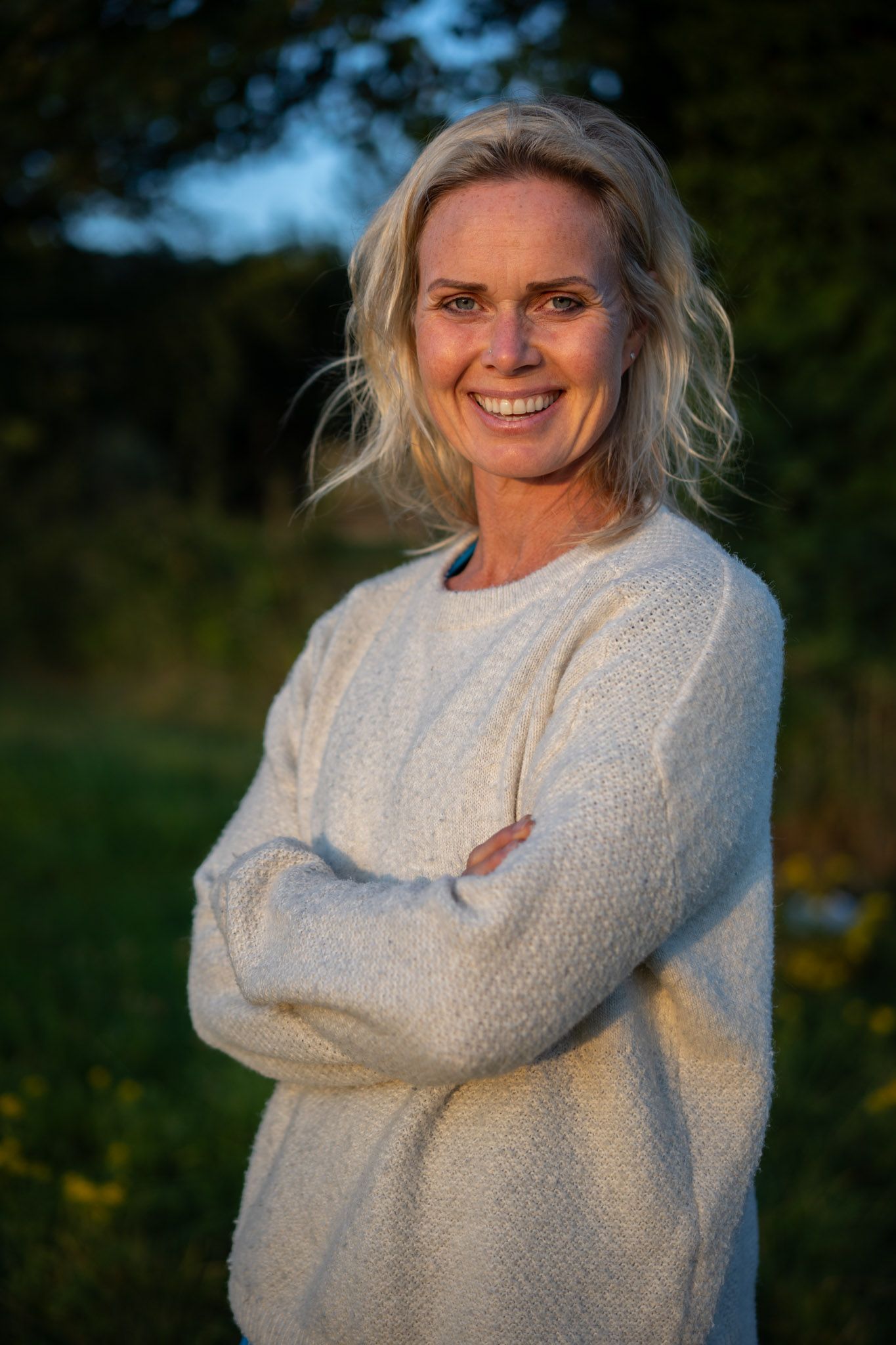 Woman smiling outdoors with arms crossed, wearing a cream sweater.