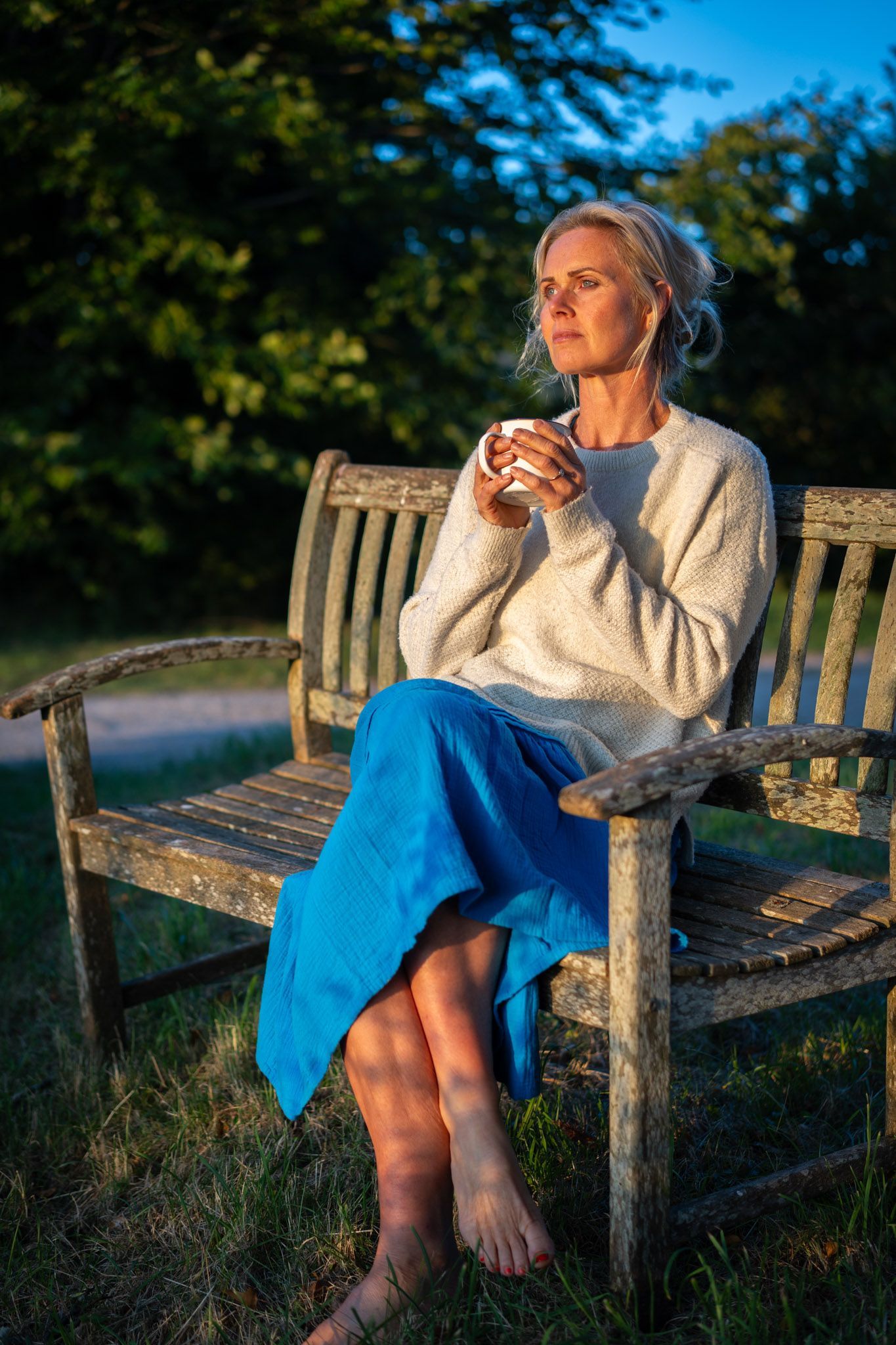 Woman sitting on a wooden bench, holding a mug. She wears a sweater and blue skirt, in a park at sunset.