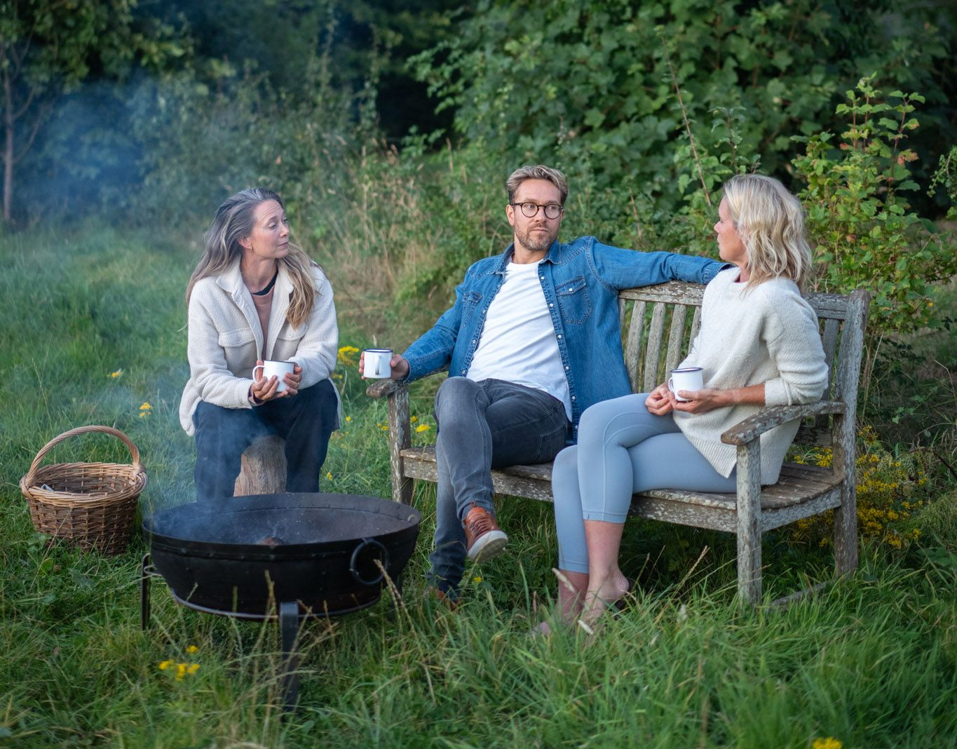 Three people around a fire pit outdoors; sitting, holding cups, and talking in a grassy area.